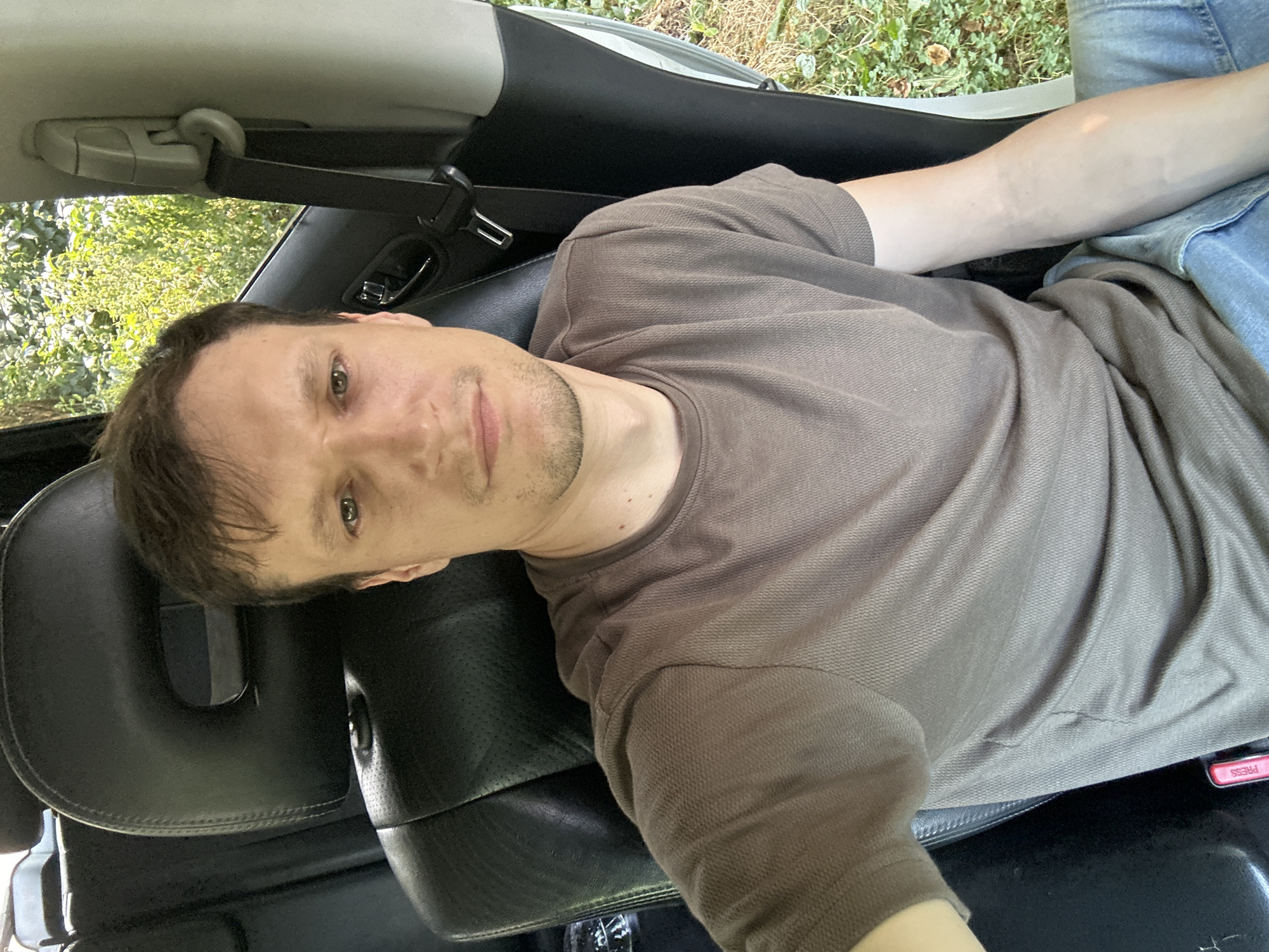 A medium shot photograph shows a man sitting in the driver's seat of a car. He is looking directly at the camera with a neutral expression. The man has short brown hair and is wearing a brown t-shirt and blue jeans. The interior of the car is visible, including the black leather seats and the seatbelt. Outside the car, trees and greenery are visible. The photograph appears to have been taken in Makiv, Ukraine.