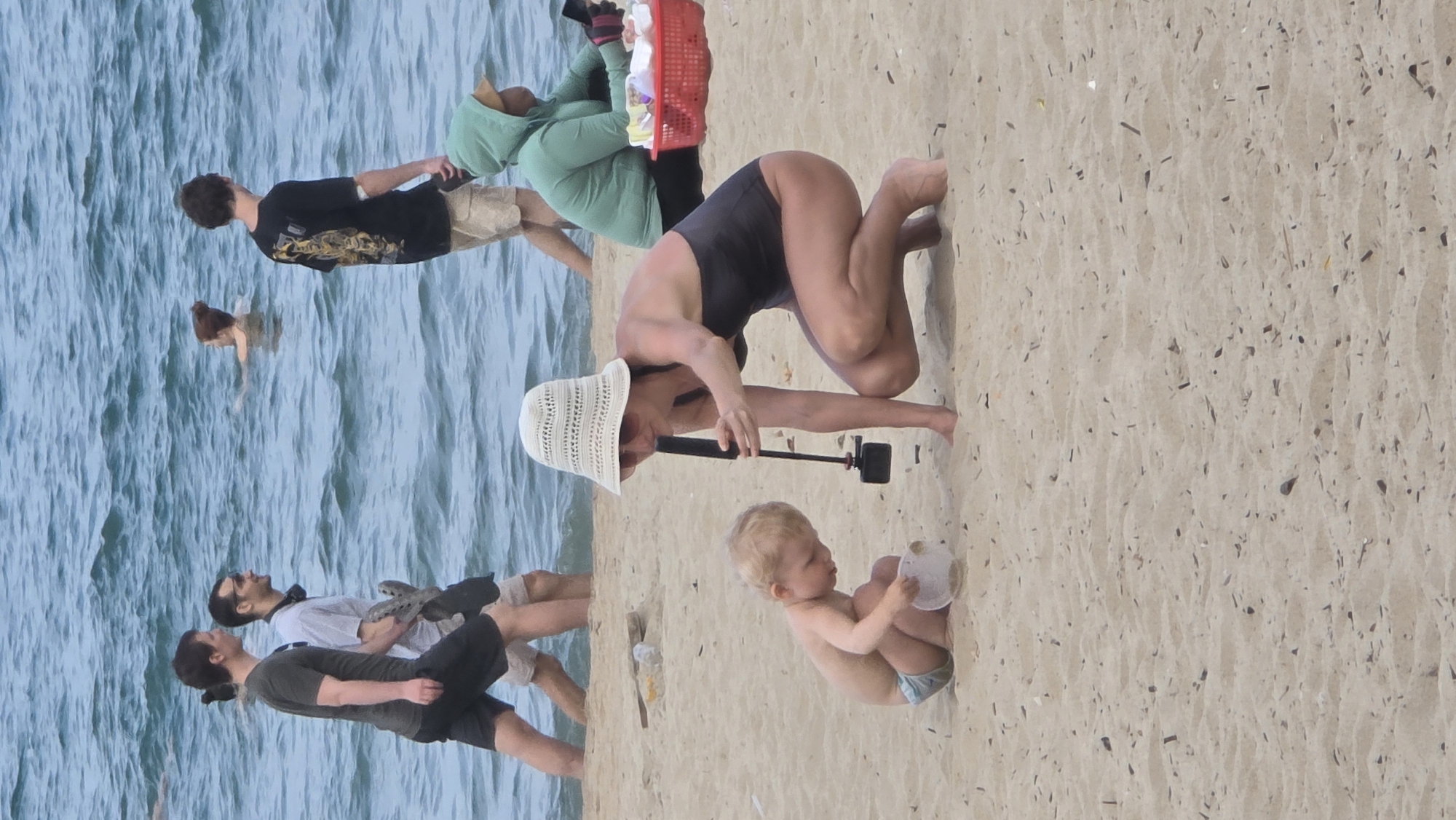 The image captures a vibrant beach scene in Nha Trang Ward, Vietnam, during daylight hours. On the right, the sandy shore is visible, populated by several individuals. A woman in a dark swimsuit and a wide-brimmed white hat is crouching on the sand, holding a camera on a selfie stick, seemingly filming a young baby. The baby, clad in a light blue diaper, sits on the sand holding a white, rounded object, possibly a toy or part of a coconut.
Towards the center and left, closer to the water, a group of people are engaged in various activities. Two men are walking along the water's edge; one wears a black t-shirt and light shorts, while the other wears a white t-shirt, dark shorts, and headphones around his neck. Further back, another person in a light green hooded top is partially visible, holding a red basket. In the calm, light blue water on the far left, the head and shoulders of a person swimming are visible. The sand itself appears light-colored and somewhat coarse, with small pieces of natural debris scattered across it. The overall atmosphere is one of leisure and recreation at the beach. No visible text is present in the image.