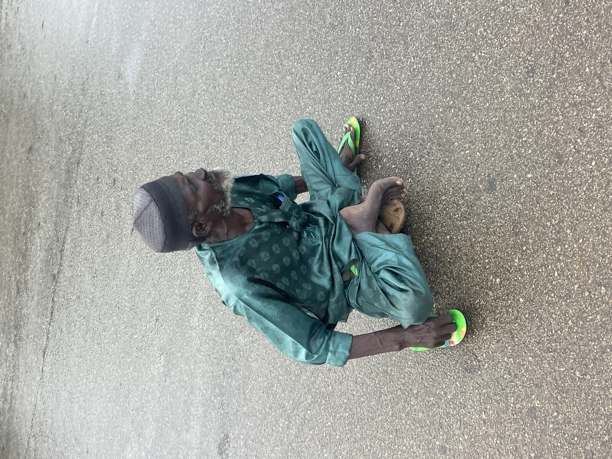 An elderly man wearing a grey cap and a teal patterned, long-sleeved tunic and pants sits cross-legged on a wet asphalt surface. The man has a light grey beard and is looking to the right. He is wearing tan sandals on his left foot and a green flip-flop on his right foot. A blue pen is visible in the chest pocket of his tunic. The scene appears to be outdoors, possibly on a street or pathway, given the wet asphalt. There are no other individuals or objects of note in the immediate vicinity. The overall impression is of a solitary figure in an urban environment.
