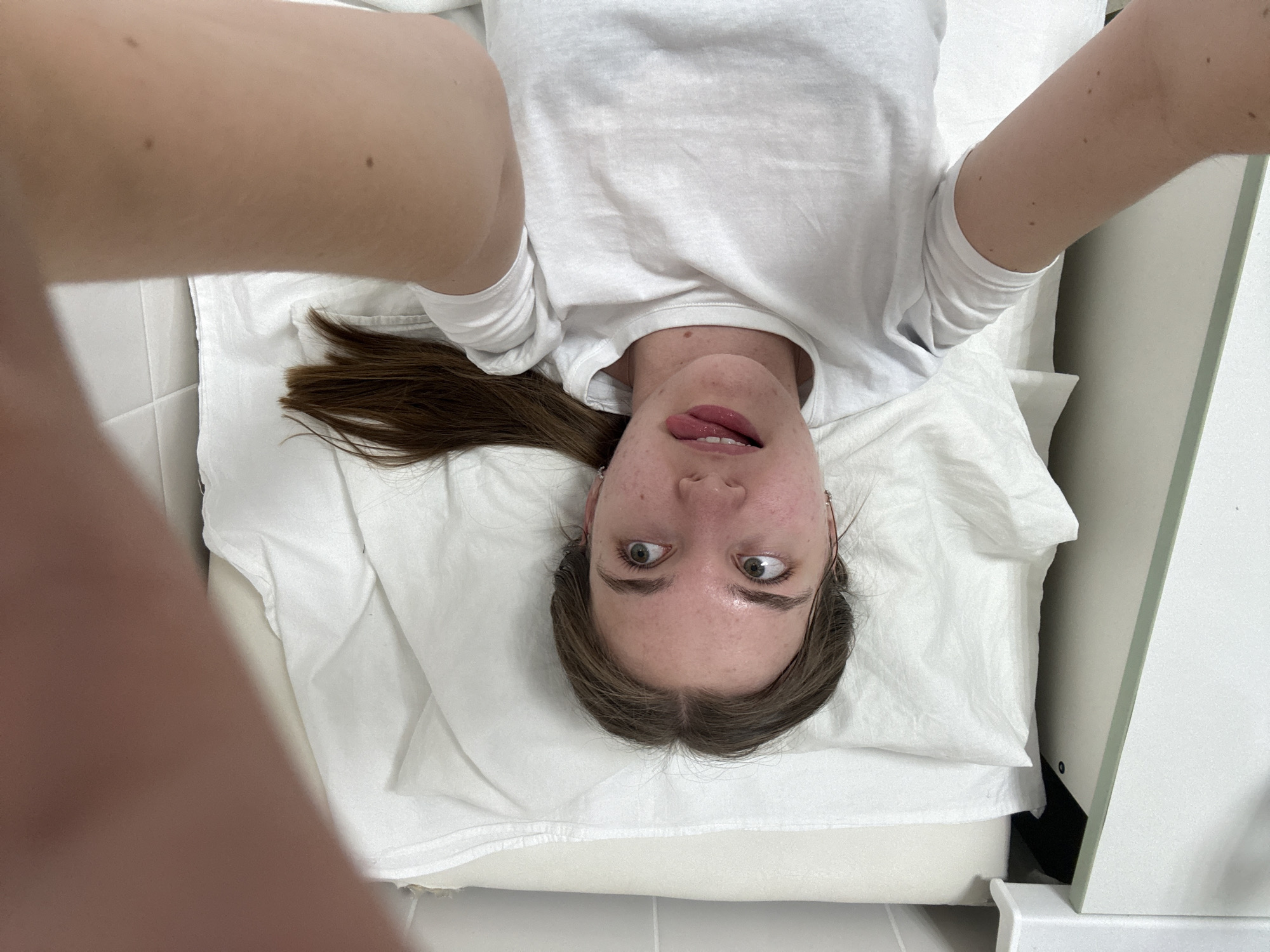 A woman with light brown hair is lying on a white examination table, likely in a medical office or clinic. The woman is wearing a white t-shirt. She has her arms raised, possibly taking a selfie. Her tongue is sticking out slightly. She is Caucasian. The location is likely in Perm, Russia.