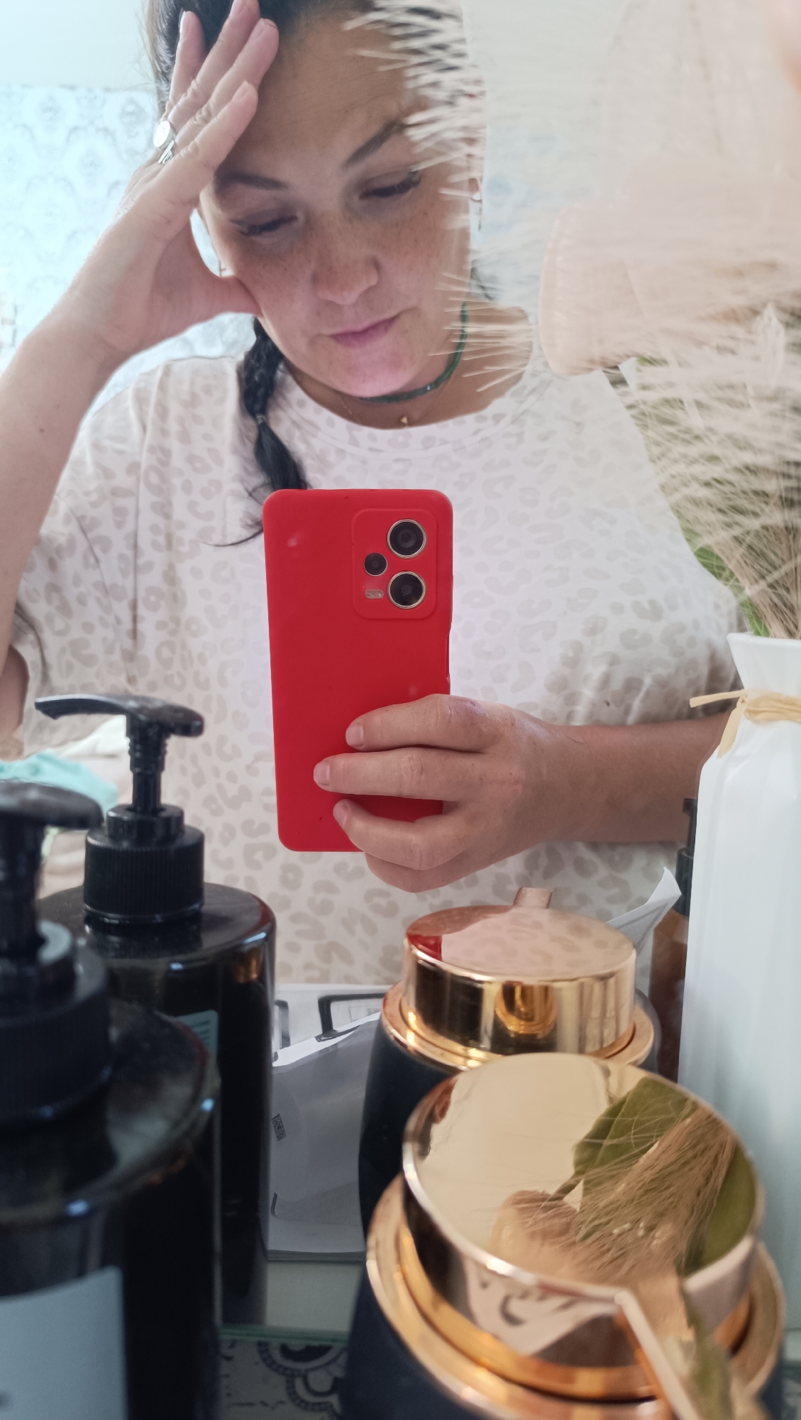 The image shows a woman holding a red smartphone in her right hand, taking a selfie in a mirror. She has her left hand on her forehead, and her eyes are cast downwards. She is wearing a white t-shirt with a leopard print pattern. In the foreground, out of focus, are several cosmetic bottles and jars with black and gold accents. The background appears to be a bathroom or dressing area, with tiled walls visible. The woman has freckles and her hair is styled in a braid. The image does not provide any information or visual cues that would suggest a specific location in Chaikovsky, Russia.