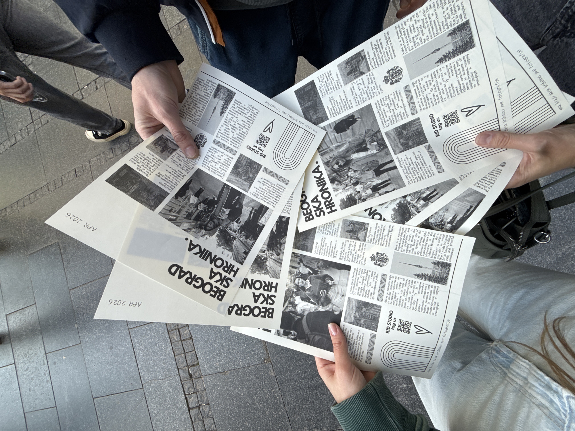 An overhead photograph captures several individuals holding and distributing multiple black and white printed papers outdoors on a paved surface. Two sets of hands are prominent in the foreground, one holding a thick stack of papers and another holding a few individual sheets. The lower bodies and legs of at least three people are visible, dressed in casual attire including jeans and sneakers, suggesting an urban setting like a sidewalk or public square. A small black bag is partially visible near one person's leg, and a mobile phone in another's hand.
The printed materials are styled like a newspaper or zine, titled "BEO GRAD SKA HRONIKA." (Belgrade Chronicle) in large, bold lettering. The date "APR 2026" is visible on the bottom corner of some sheets. The papers feature articles in both English and Serbian, accompanied by black and white photographs of Belgrade landmarks, such as the Avala Tower, and various street scenes. Specific textual references include "Strahinjića Bana Street" and "Belgrade Fortress," consistent with the Belgrade, Serbia location context.
Each sheet prominently displays "RID STUDIO," a QR code, a heart icon, and the phrase "tag us." Another notable Serbian phrase, "Na kraju dana vréđamo sve fotografije," is also visible. The activity appears to be the distribution or presentation of these local chronicles during daytime, indicated by the bright, natural light.