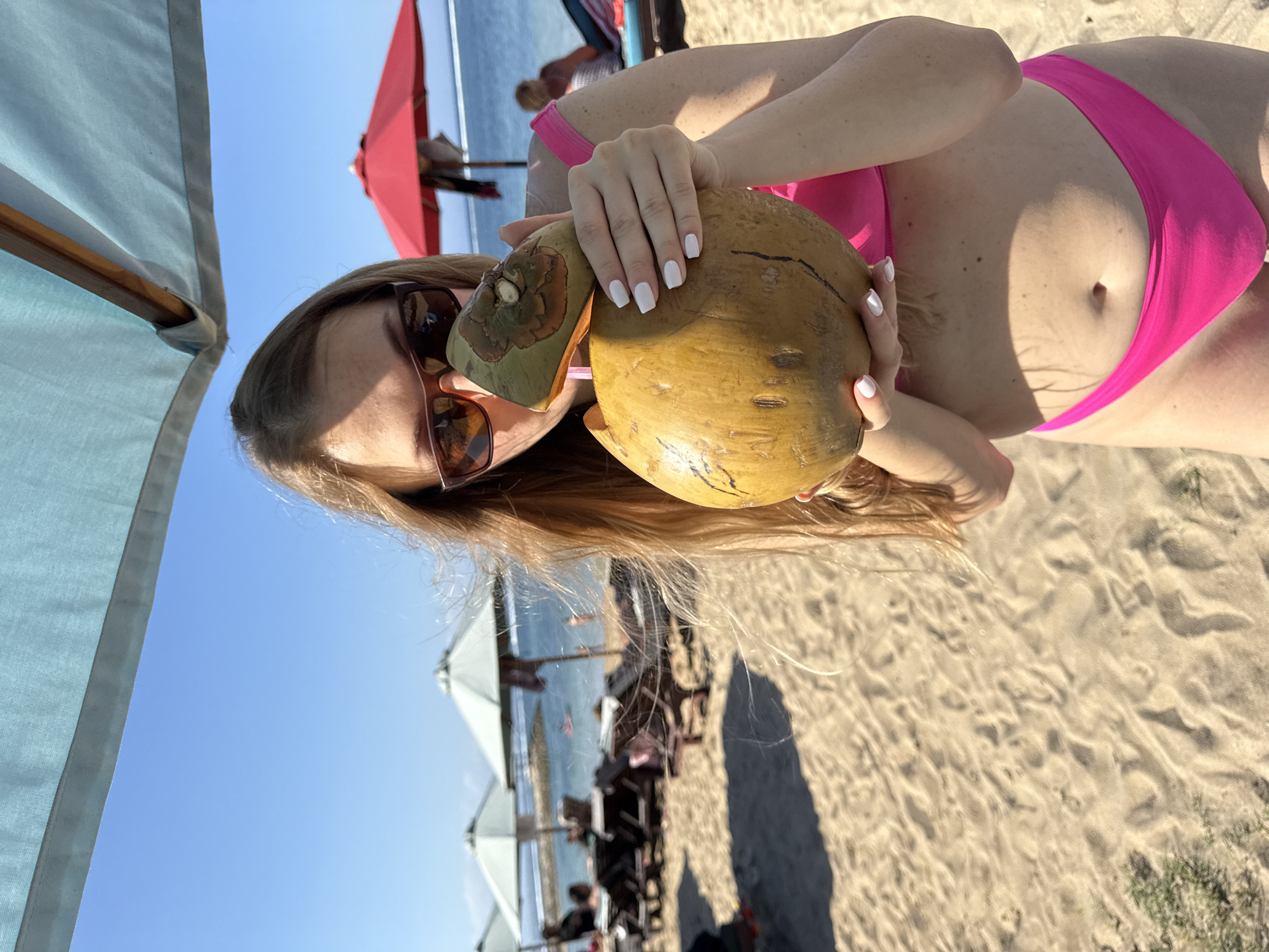 A young Caucasian woman with long blonde hair and sunglasses is holding a young coconut and drinking from it on a sandy beach. She is wearing a pink bikini. The beach has lounge chairs and umbrellas in the background, with the ocean visible beyond them. The sky is clear and blue. The location appears to be a beach resort area within Denpasar, Indonesia, likely on the coast of Sanur or Nusa Dua given the typical amenities shown.