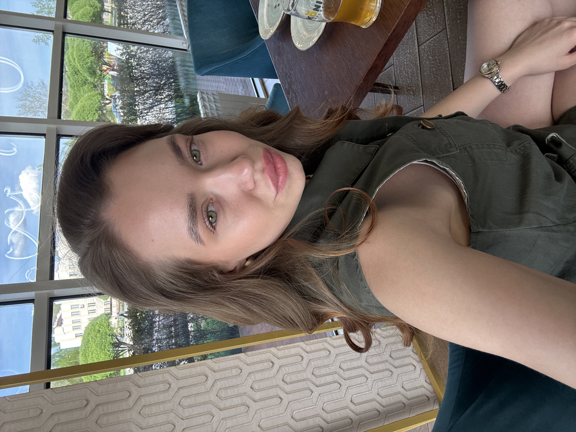 A woman is seated in an indoor setting, likely a restaurant or cafe, in what appears to be Chaikovsky, Russia. The exterior of a building can be seen through a multi-paned window, along with trees in the background. The woman is wearing an olive green shirt. She also has a wristwatch on her wrist. A table next to her holds a beer glass, and another turquoise seat can be seen next to her. The table has two dishes on it.