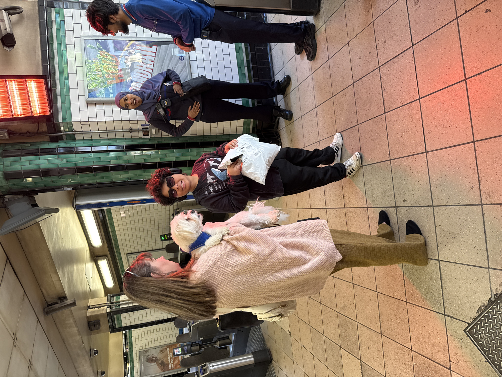 The image depicts the interior of a London Underground station, likely a platform or concourse area. The floor is tiled in a light beige color. A row of automated ticket barriers is visible. There are several people present. A woman with long brown hair and a light pink sweater is holding a small white dog with a blue collar. Behind her, another person is sitting with a white package in their lap. In the background, two other people are standing. There's a red heater on the wall, as well as tiled walls with green and white sections. The lighting is a mix of fluorescent and warm tones. There are advertisements or informational posters on the walls.
