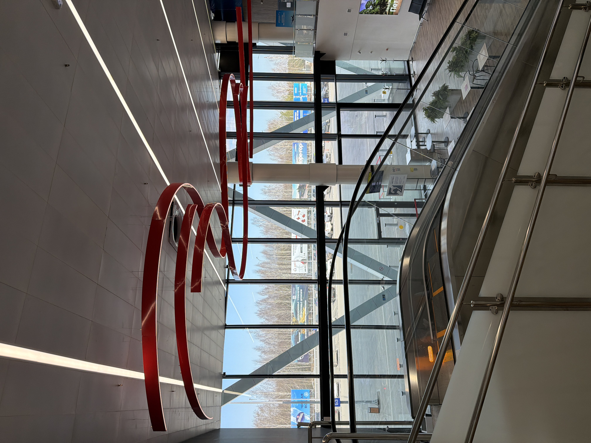 This media file captures a bright, modern indoor space, likely a public building such as an airport terminal or a large commercial center in Izhevsk, Russia. The image appears to be taken from an upper level, looking across a wide hall towards large windows and an ascending escalator.
On the left, a tall, light-colored wall features sleek horizontal strip lighting and a prominent, abstract red sculpture. The sculpture is made of curved, ribbon-like elements that extend across a significant portion of the wall, creating a dynamic visual focal point.
The center of the frame is dominated by a multi-pane glass facade, revealing the exterior. Beyond the windows, the scene shows a clear daytime sky over an outdoor area with numerous bare trees, suggesting a late autumn or winter season. A road or parking area is visible, along with distant buildings and several large billboards displaying Cyrillic text. Notable text includes "ИННОВАЦИОННЫЕ ТЕХНОЛОГИИ ПРОЧНЕЙШИЕ АРТИТЕЛИ" (Innovative Technologies Strongest Artels), "ОТКРЫВА" (Open...), and partially visible phrases such as "чи ая во" (possibly part of "Чистая вода" - Clean Water) on a blue sign, and another blue sign with "54".
To the right, an escalator with glass side panels and polished stainless steel handrails ascends towards an upper floor. Through the glass barrier of the escalator, a lower level is visible, furnished with modern tables, chairs, and potted plants, suggesting a cafe or lounge area. No people are visible, indicating a quiet moment in this public space. The interior is well-lit by both the artificial strip lights and abundant natural daylight. A partial sign on the upper right of the interior reads "ПЕТР".