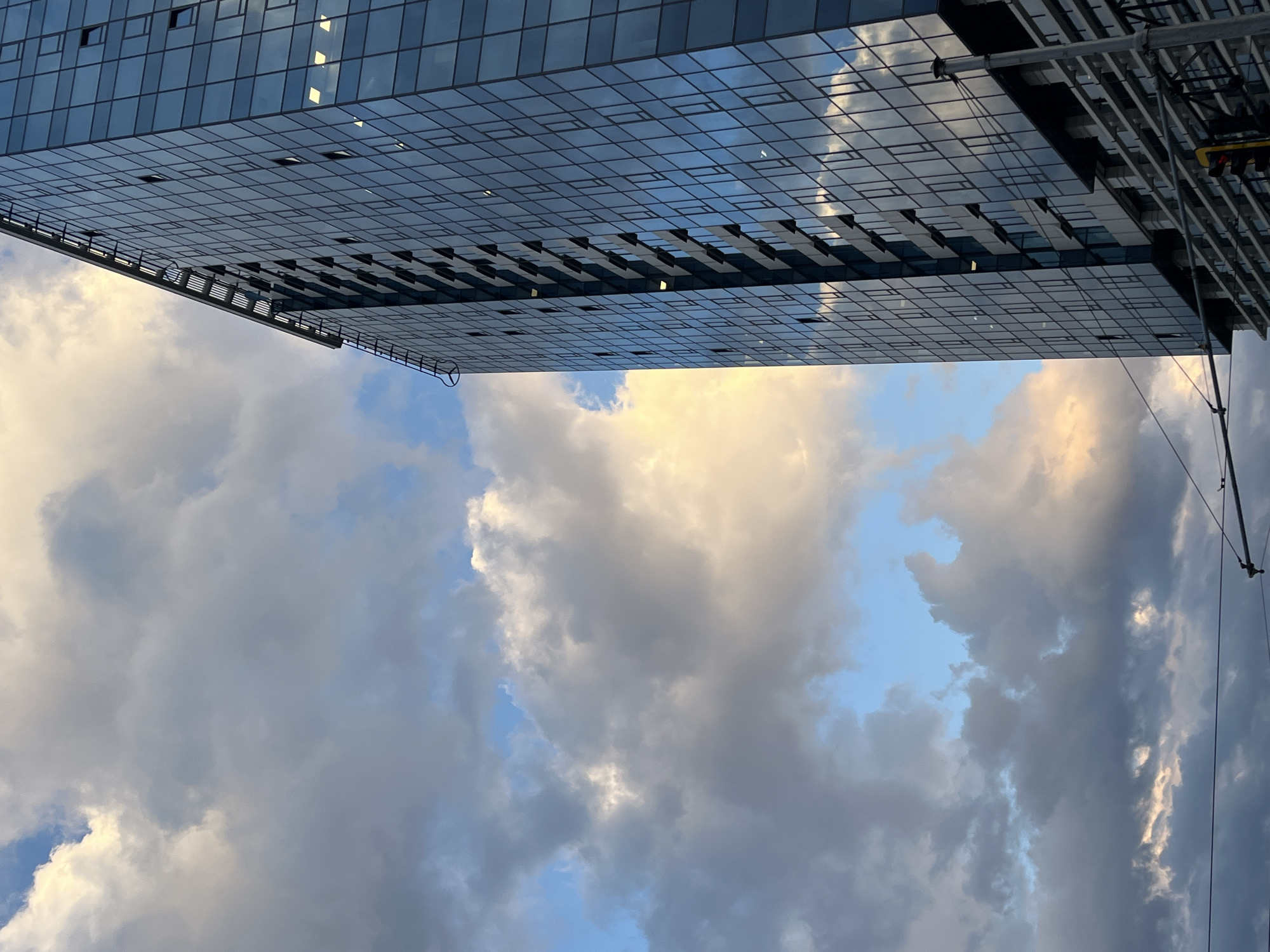 An image captures a modern glass-paneled building against a dramatic sky in Sofia, Bulgaria. The building, a multi-story structure characterized by its extensive grid facade of reflective windows, occupies the upper portion of the frame, extending from the top left towards the bottom right. This perspective suggests a view looking upwards at an angled or possibly cantilevered section of the architecture. The reflective surface of the building mirrors the cloudy sky, and some windows reveal interior illumination. Along the lower edge of the building, distinct structural elements resembling maintenance rings or small ladders are visible.
The lower part of the image is dominated by a dynamic sky, filled with textured clouds that display a wide range of tones. These clouds transition from bright whites and warm golden hues, where they are illuminated by sunlight, to darker shades of grey and blue. Patches of clear blue sky are intermittently visible between the cloud formations. The lighting suggests either a late afternoon or early morning, creating a striking contrast between the cool, architectural lines of the building and the vibrant, ever-changing sky. On the far right edge, elements of urban infrastructure, including thin wires and a metal support structure (possibly for a traffic light), are partially visible, confirming an urban setting. No text is visible in the image.