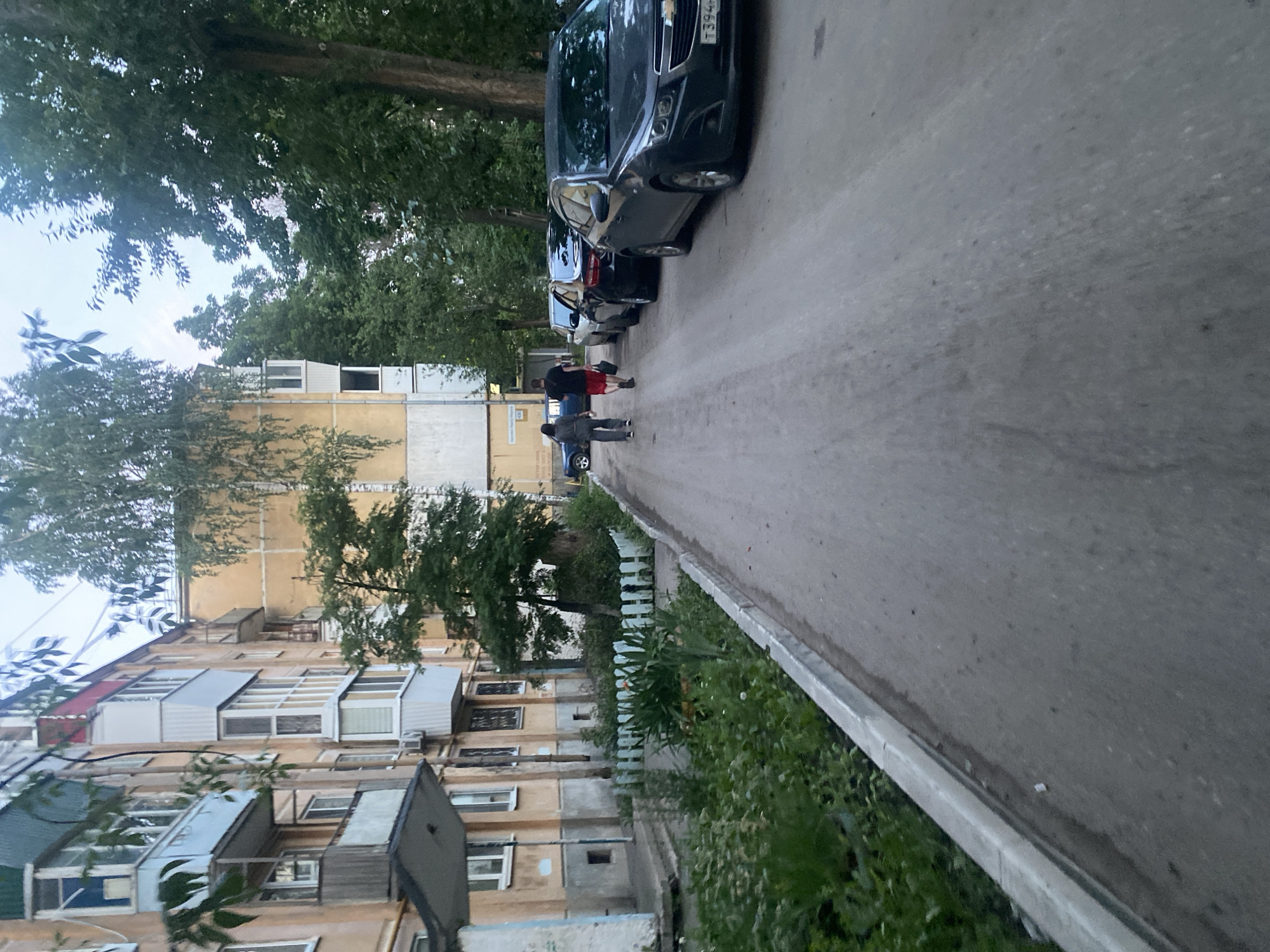 The image shows an outdoor scene in front of an apartment building in Samara, Russia. A paved pathway runs alongside a grassy area with some fencing and plants. Several cars are parked along the pathway. Two individuals are walking away from the camera on the pathway. The apartment building appears to be old, with a light yellow facade and balconies. Trees are visible in the background.