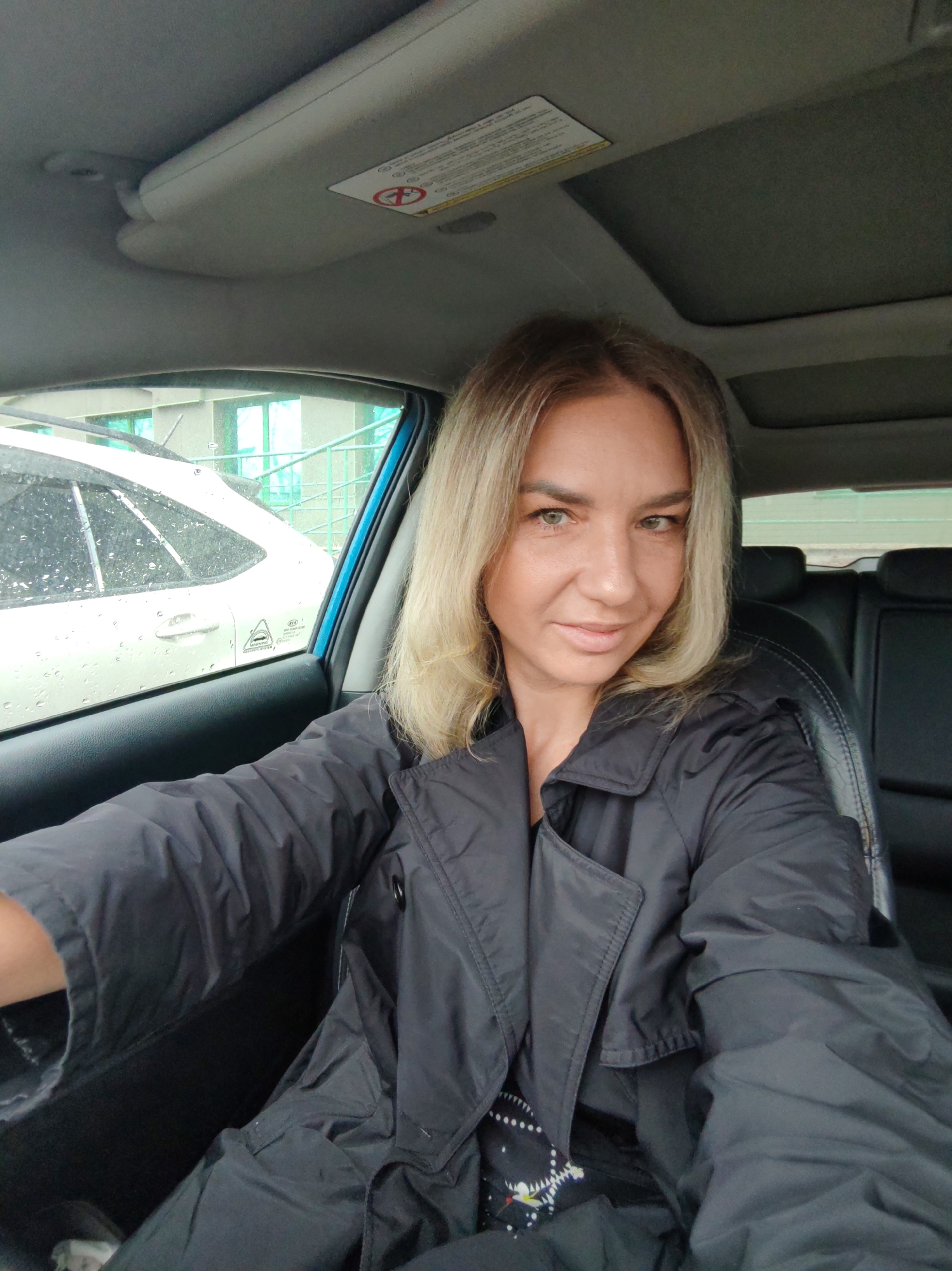 A woman is in a car, possibly the driver's seat. The car is likely parked or stopped, given the indoor setting. Outside the window, another car is visible, and a building with light green accents can be seen. The woman is wearing a dark jacket, and her hair is blonde. The car's interior appears to be mostly black. The location is unspecified, but the prompt mentions Petropavlovsk-Kamchatsky, Russia. It is raining outside.