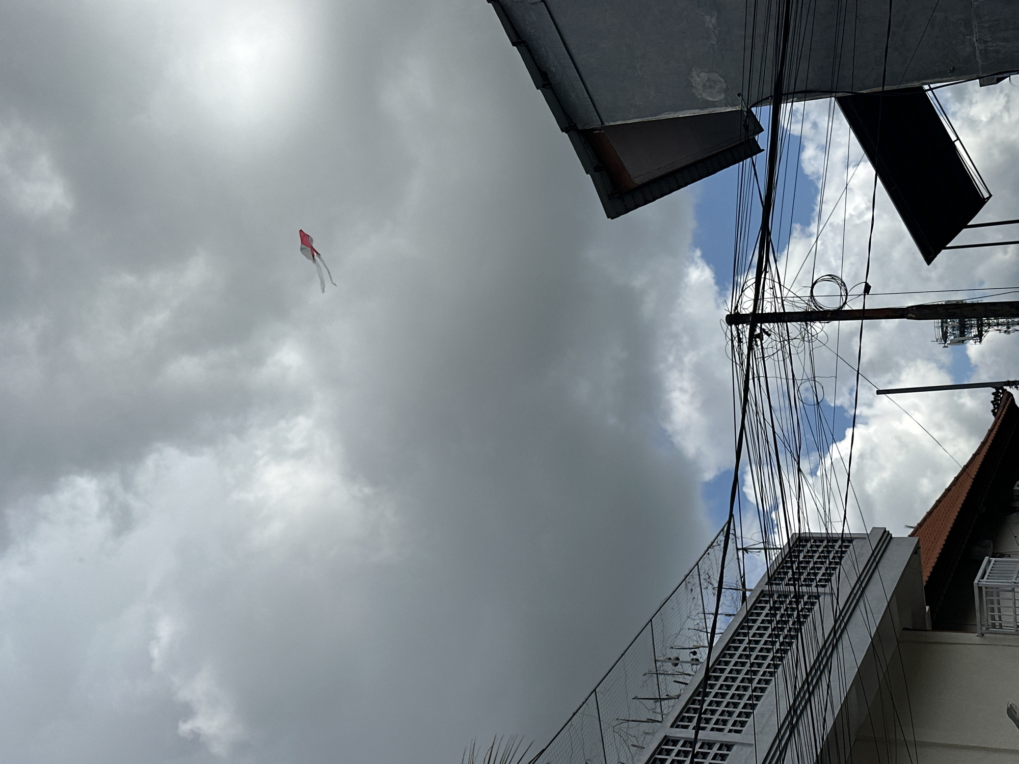 The image shows a cloudy sky above the city of Dalung, Indonesia. A kite with a red and white pattern is flying in the distance. The view is from a low angle, looking up at buildings, roofs, and a network of electrical wires. A staircase with a patterned facade is visible against the backdrop of clouds. The weather appears overcast.