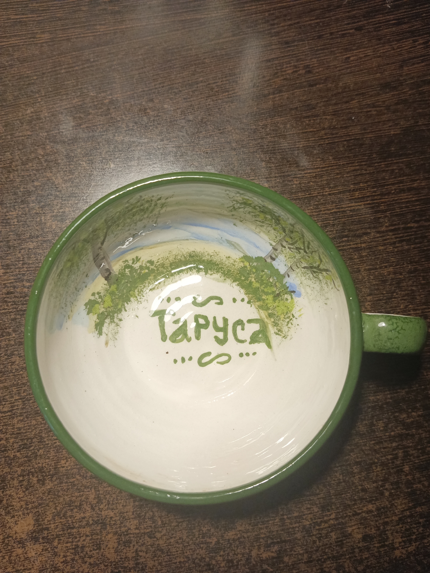 The image shows a ceramic bowl with a green rim. The inside of the bowl is white and decorated with a painted landscape featuring trees and a body of water. The word "Таруса" (Tarusa) is written in green lettering in the center of the bowl, with decorative dots above and below it. The bowl is placed on a dark wooden surface, likely a table. The scene is an indoor setting, and the lighting suggests it is either dimly lit or the flash from the camera is the primary light source. There are no people visible in the image. The bowl appears to be a souvenir or decorative item, possibly depicting a scene from the city of Tarusa, Russia. The image does not provide any specific information about the city of Obninsk, Russia.