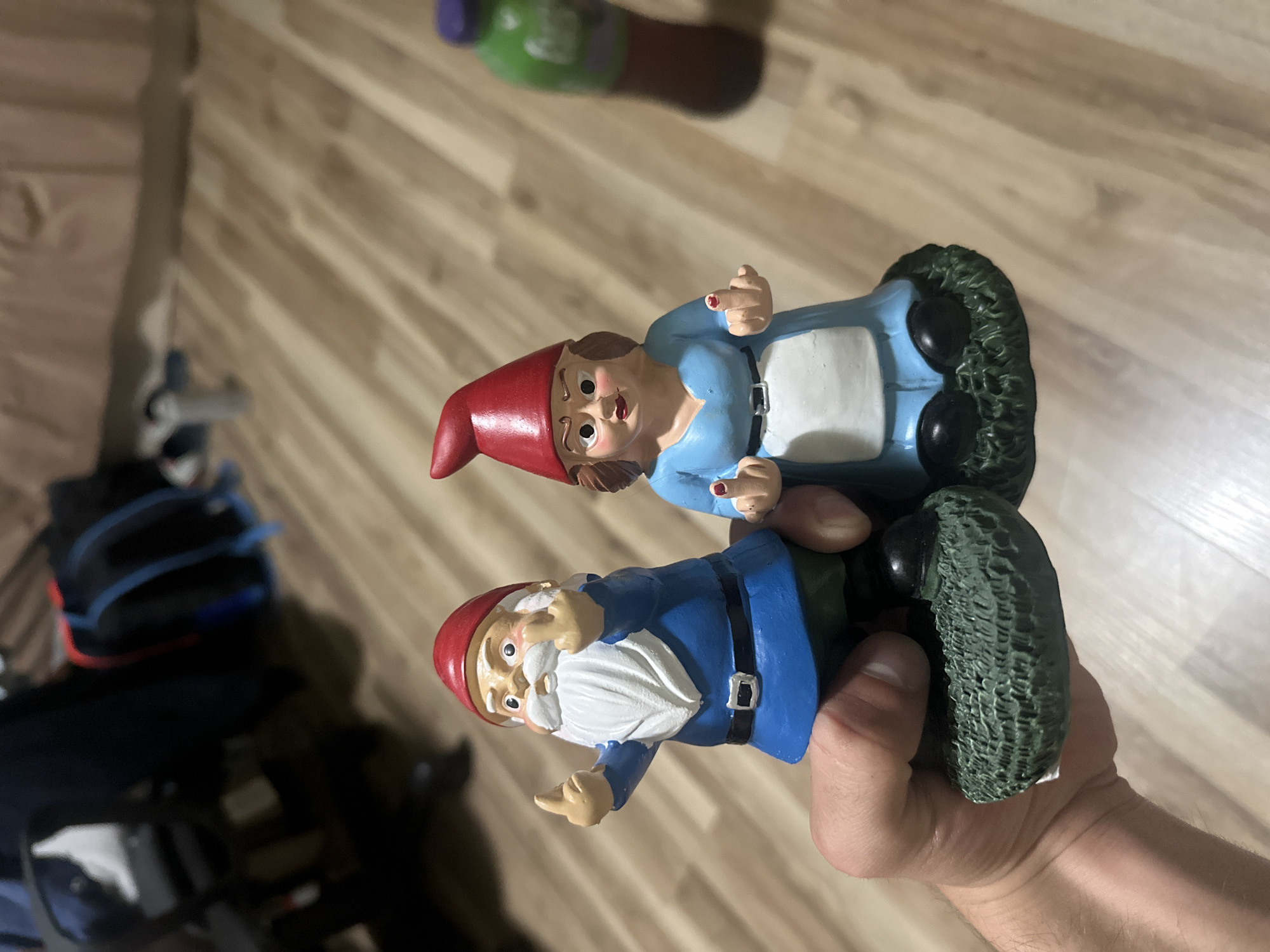 The image depicts two garden gnomes, a male and a female, displaying offensive hand gestures (middle fingers). The male gnome, wearing a blue tunic, black belt, white beard, and red conical hat, has an angry expression. The female gnome, dressed in a light blue outfit with a white apron and a red conical hat, also has an angry expression, with visible red nail polish on her raised middle fingers. A human hand, positioned in the lower-left, grasps the base of the male gnome. Both gnomes stand on green textured bases. The setting is an indoor location in Stara Zagora, Bulgaria, featuring a light brown laminate wood floor. The blurred background elements include a dark office chair, a blue and red backpack, and a green and purple plastic bottle. A light brown fabric is visible in the distant background.