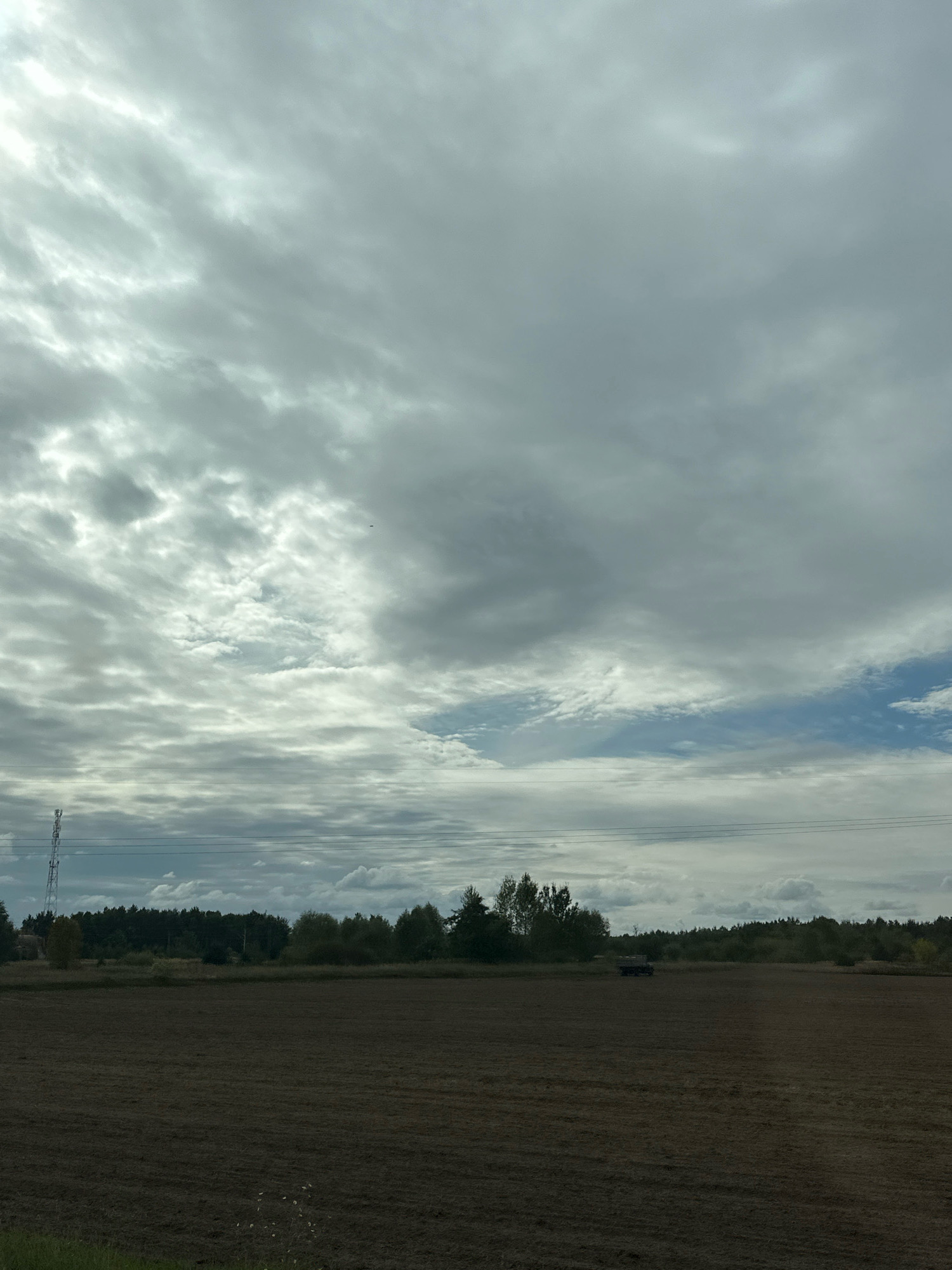 The image depicts a wide, open field, likely a recently harvested or prepared agricultural plot, under a dramatic, overcast sky. The foreground is dominated by the dark, tilled earth of the field, showing distinct rows or patterns from agricultural machinery. In the middle ground, a line of trees forms a dense, dark green border against the horizon. To the left, a tall, lattice-like telecommunications tower is visible, and further into the field, a dark-colored vehicle, possibly a tractor or utility vehicle, is parked or in motion. Power lines stretch horizontally across the scene, connecting to the tower and extending towards the right. The sky is filled with layers of gray and white clouds, with some patches of brighter sky and sunlight breaking through, creating a dynamic and diffused lighting condition. The overall impression is one of a rural, working landscape under a changeable sky. The location is identified as Парыцкі сельскі Савет, Belarus.