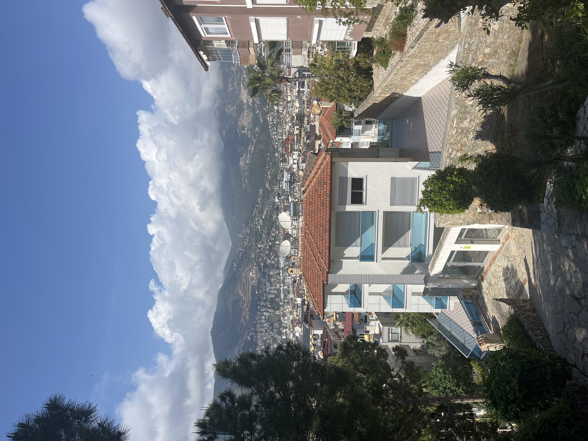 The image presents a panoramic view of a coastal city, likely Alanya, Turkey, from an elevated perspective. The scene is dominated by urban development cascading down a mountainside, framed by a clear blue sky with large, fluffy white clouds.
In the foreground, parts of several buildings are visible, including a prominent white multi-story building with distinctive blue-tinted, rectangular windows and a red-tiled roof, and a taller, brown building with white-framed windows. Lush green trees and bushes, including a palm tree, are scattered around these structures and line stone walls that define pathways and terraces. A patterned tiled courtyard or patio area is visible between some of the foreground buildings.
The midground reveals a dense cityscape extending across the lower slopes of the mountain. Numerous light-colored residential and commercial buildings with varied roof colors (many red-tiled) are packed together. Satellite dishes are visible on many rooftops.
The background features towering, rugged mountains, partially obscured by the urban sprawl and a soft haze. Large cumulus clouds hug the upper slopes of the mountains and extend across the bright blue sky.
The time of day appears to be sunny midday or early afternoon, characterized by strong sunlight and clear visibility. The weather is pleasant with clear skies and picturesque clouds. No people or specific activities are discernible in the static landscape. A small yellow sign is visible on an entrance of the white building in the foreground, but the text is unreadable.
