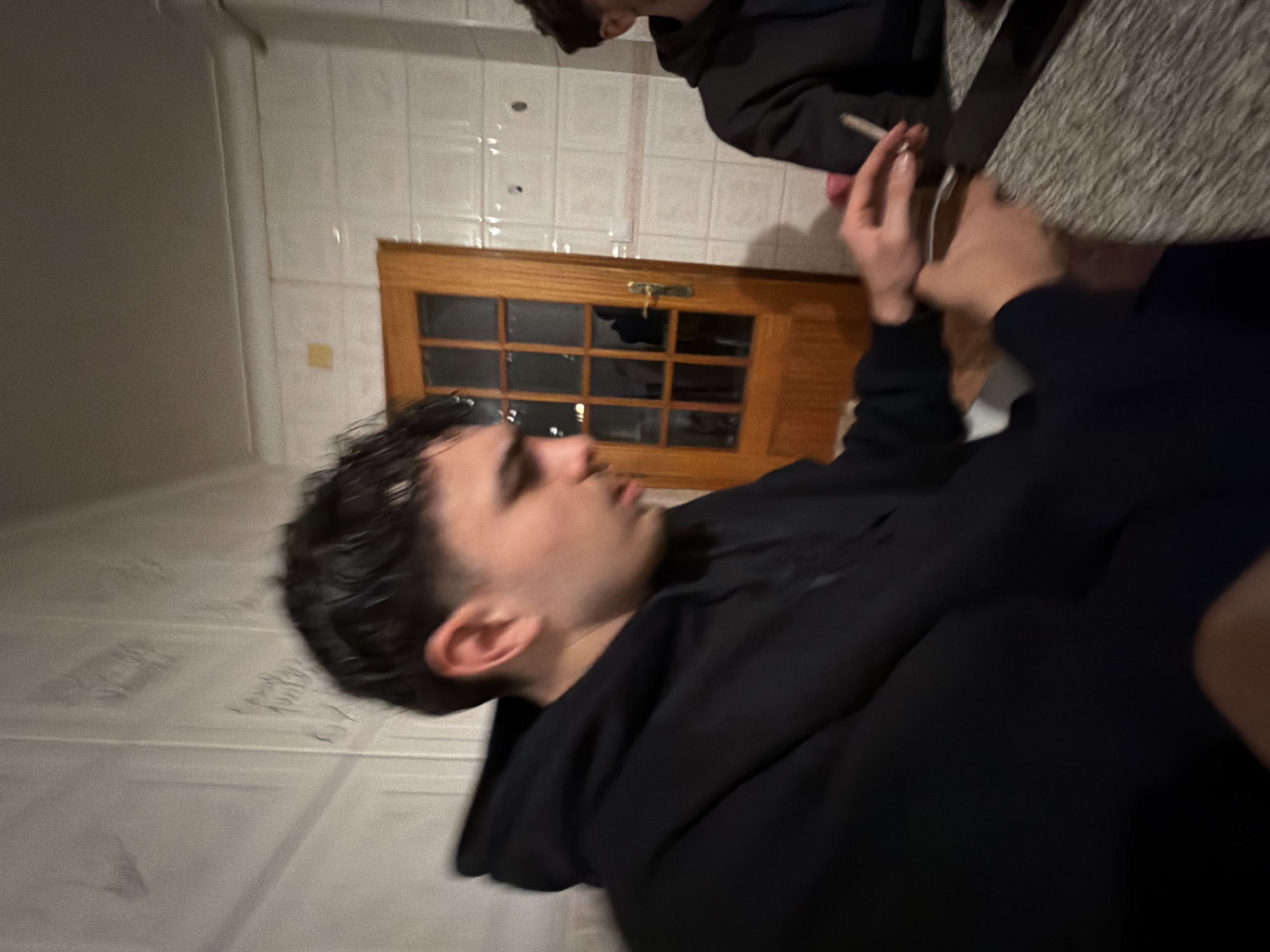 The scene is an indoor setting in Bragança, Portugal, likely a kitchen or common area, though the precise location within the city is not visually identifiable. Two young males are seated at a textured, dark-colored table. The central figure on the left, with dark hair, is wearing a black hooded sweatshirt and is looking downwards, his left hand engaging with a small dark object. A second male, partially visible on the right and also in a black hooded sweatshirt, holds a lit cigarette or hand-rolled cigarette in his right hand. The background consists of white, glazed ceramic tiled walls with visible grout lines and faint inscribed markings. A light-brown wooden door with multiple glass panes and a brass-colored handle is positioned behind the figures, reflecting a faint image of a person within its glass.