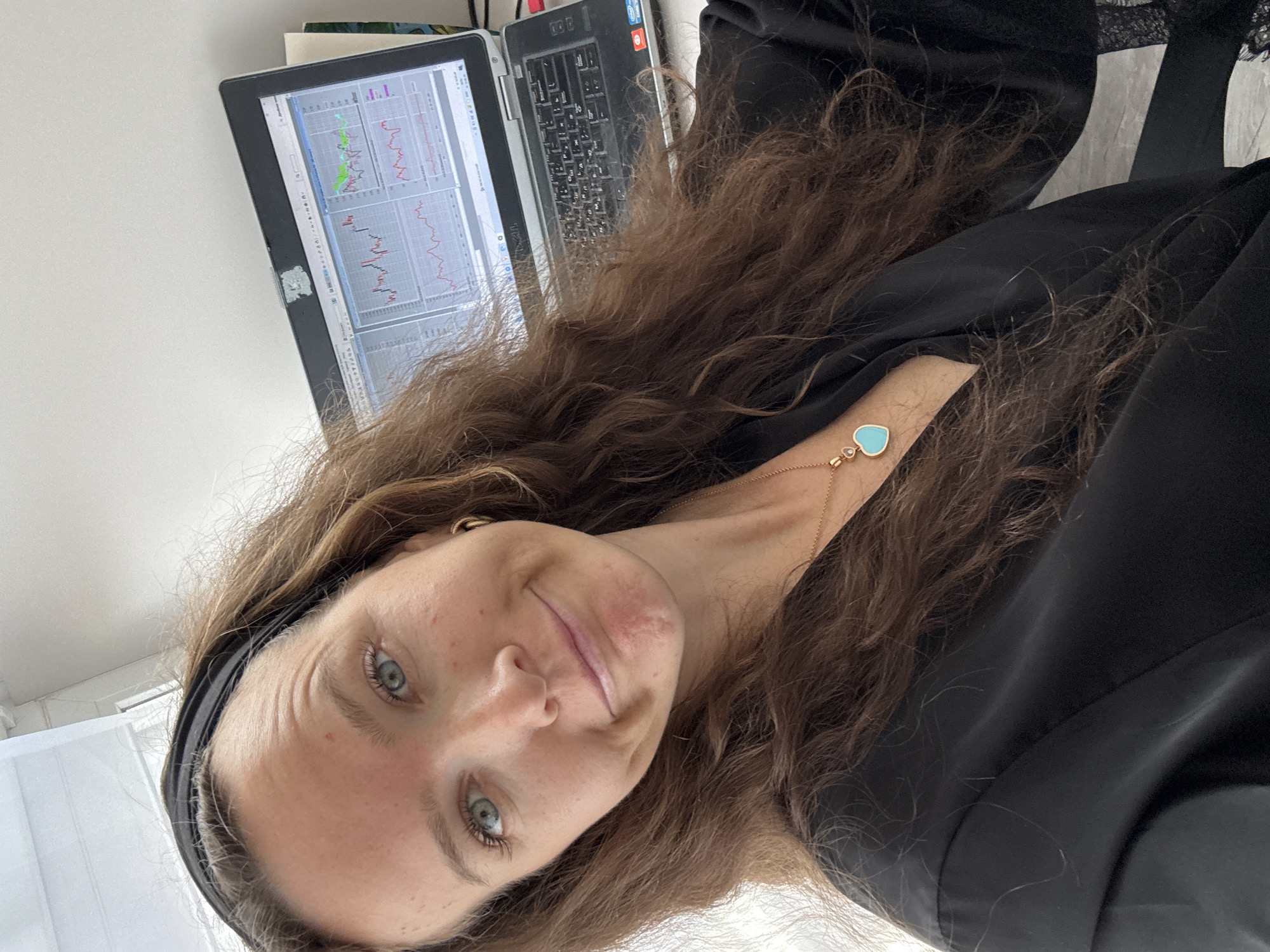 A medium shot captures a woman with long brown hair and blue eyes looking directly at the camera, her head tilted slightly and a subtle smile on her face. She is wearing a black top and a necklace with a turquoise heart pendant. A black headband is visible on her head. In the background, a laptop displays financial charts. The scene appears to be an indoor setting, possibly an office or home workspace, with a window and white walls visible. The image is taken from a slightly upward angle.