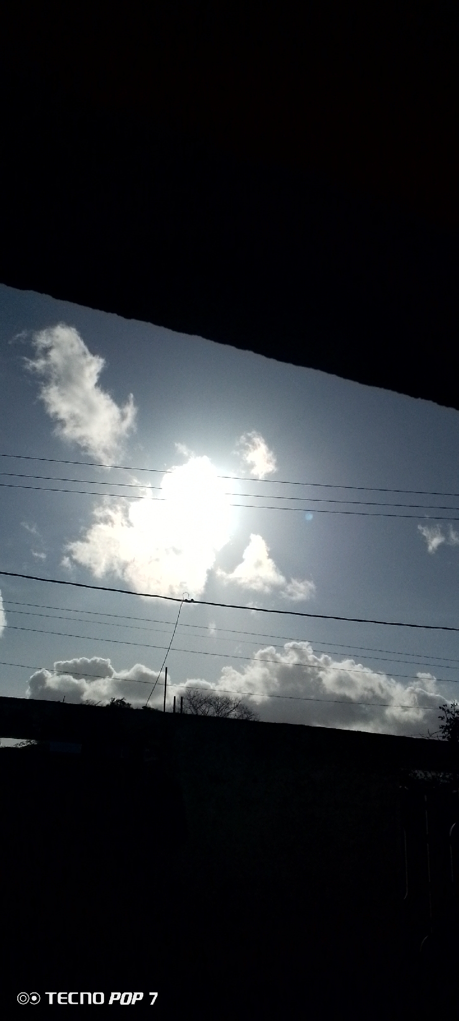The image depicts a view of the sky with clouds and the sun, framed by dark, silhouetted structures at the top and bottom. Three electrical wires stretch horizontally across the middle portion of the image. The sun is positioned near the center, obscured by white clouds. A pole with a thin wire extending from it is visible. The lower portion of the frame contains an indistinct landscape of trees. The image includes the text "TECNO POP 7" in the bottom left corner. The precise location in Altagracia, Venezuela is impossible to determine from this single image.