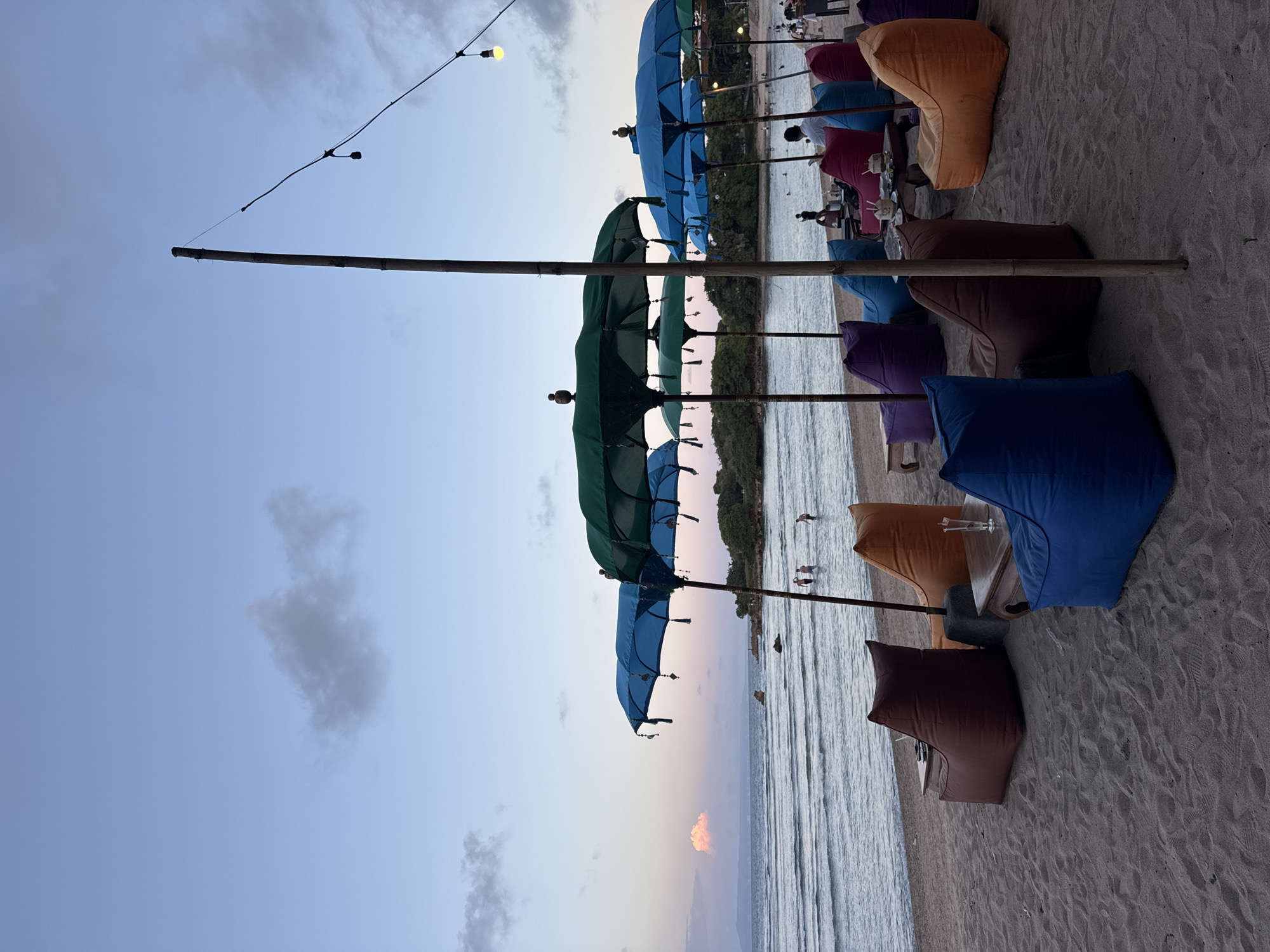 The image shows a beach scene at sunset in an unidentified location in Indonesia. The foreground features colorful beanbag chairs and low tables arranged on the sand. Several rows of open umbrellas with bamboo poles extend into the background, providing shade. Beyond the umbrellas, the ocean is visible, with a few people wading in the water. The sky is a mix of blue and pink hues, indicating sunset. String lights are strung above the umbrellas, suggesting a restaurant or lounge setting. The overall impression is of a relaxed, beachside atmosphere.