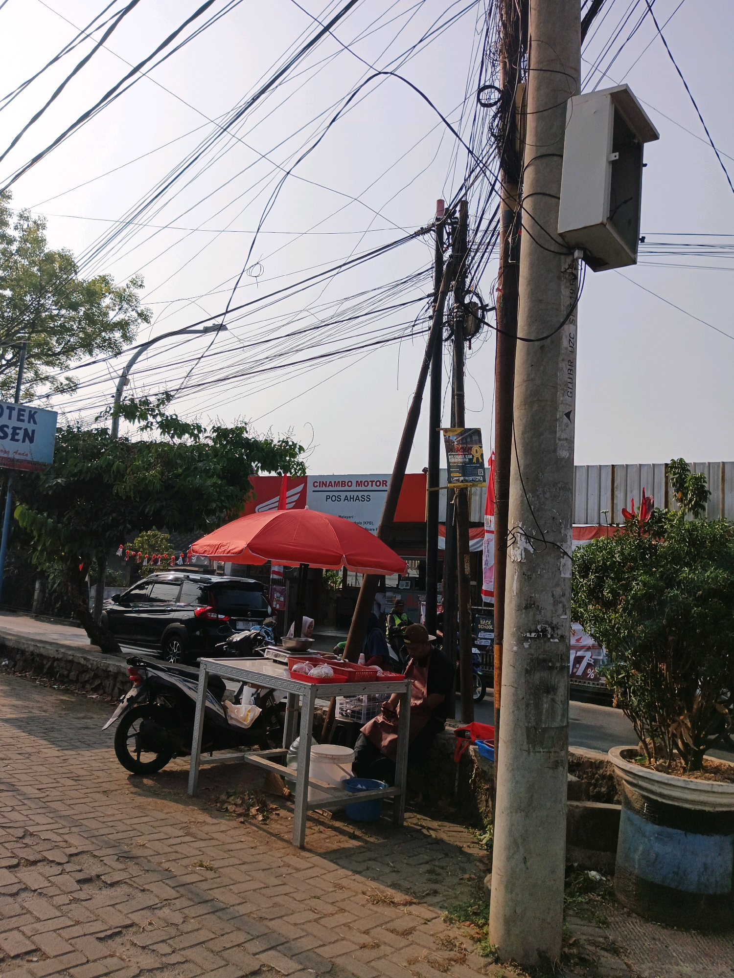 The image captures a street scene in Bandung City, Indonesia, characterized by a dense network of overhead electrical wires crisscrossing the frame against a pale sky. In the foreground, a paved sidewalk made of interlocking bricks leads to a roadside stall. A red umbrella shades a table laden with items, possibly food, and a motorcycle is parked beside it. To the left, a black SUV is parked on the street. In the background, trees and buildings are visible, including signage that reads "OTEK ASEN" and "CINAMBO MOTOR POS AHASS". A person is seated near the stall, appearing to be interacting with the items on the table. A concrete utility pole with an attached white box is prominently featured on the right side of the image. The overall impression is of a bustling urban environment with a mix of commercial activity and everyday life.