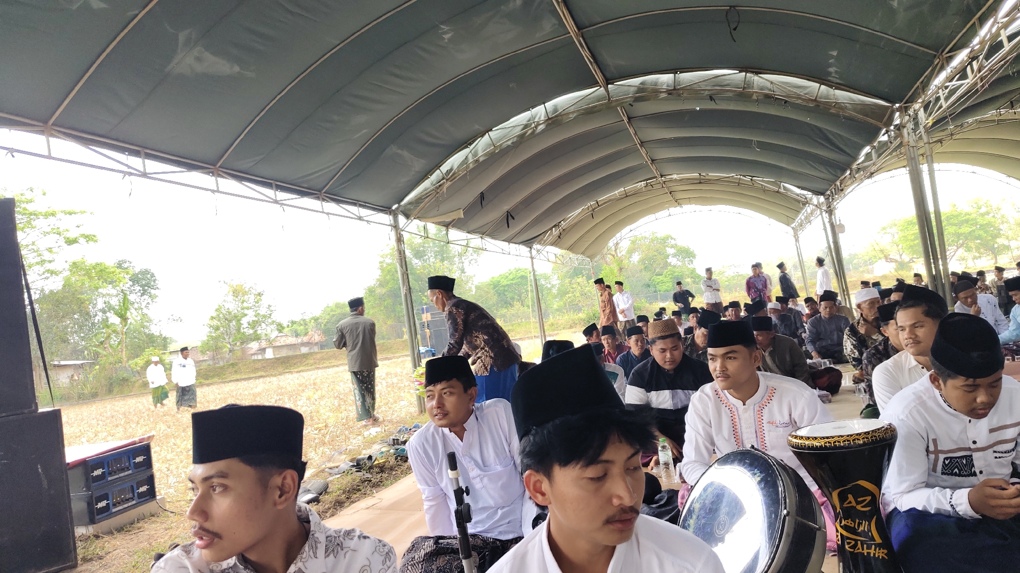 In Telagah, Indonesia, a group of men, mostly young, are gathered outdoors under a large, dark green canopy supported by metal beams. The setting appears to be rural, with dry, harvested fields visible in the background, interspersed with trees and some simple buildings. The sky is overcast and bright.
The men are dressed in traditional Islamic attire, predominantly white shirts and peci (black caps). Several individuals are seated on the ground, while others stand. Some men are in the foreground, looking towards the camera or engaged in conversation. One young man in the immediate foreground wears a white shirt with intricate embroidery and a black peci. To his right, another young man in a white shirt and black peci is seated.
In the midground, men are seated in rows, listening to something or observing. A man in a patterned brown and black tunic is bending down, perhaps interacting with someone or an object. Further back, more people are visible, some standing and others seated. Two men in white shirts and peci stand together in the mid-left of the frame.
Objects present include large speakers to the left of the frame, and two drums, one black with gold Arabic lettering and the other silver and metallic, are prominently placed in the foreground, suggesting a possible musical or religious event. A water bottle is visible on the ground. The ground itself is a mix of dry earth and some scattered debris. The overall atmosphere seems to be one of a communal gathering.