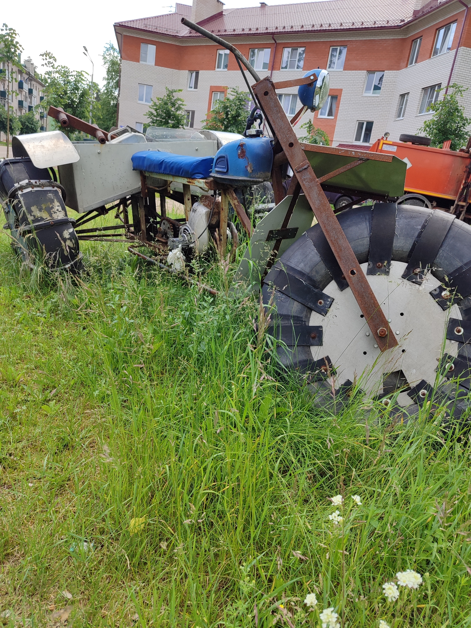 An abandoned, rusty, homemade amphibious vehicle sits partially submerged in tall green grass in an urban setting. The vehicle is primarily composed of metal parts, including a light-colored frame, a blue seat, and large, unusual wheels with segmented rubber treads. A blue fuel tank and a single blue headlight are visible at the front. In the background, a multi-story brick apartment building with a red roof stands under an overcast sky. Other assorted debris and foliage surround the vehicle. The scene appears to be in a neglected outdoor area, possibly a vacant lot or the edge of a park, within a city.
