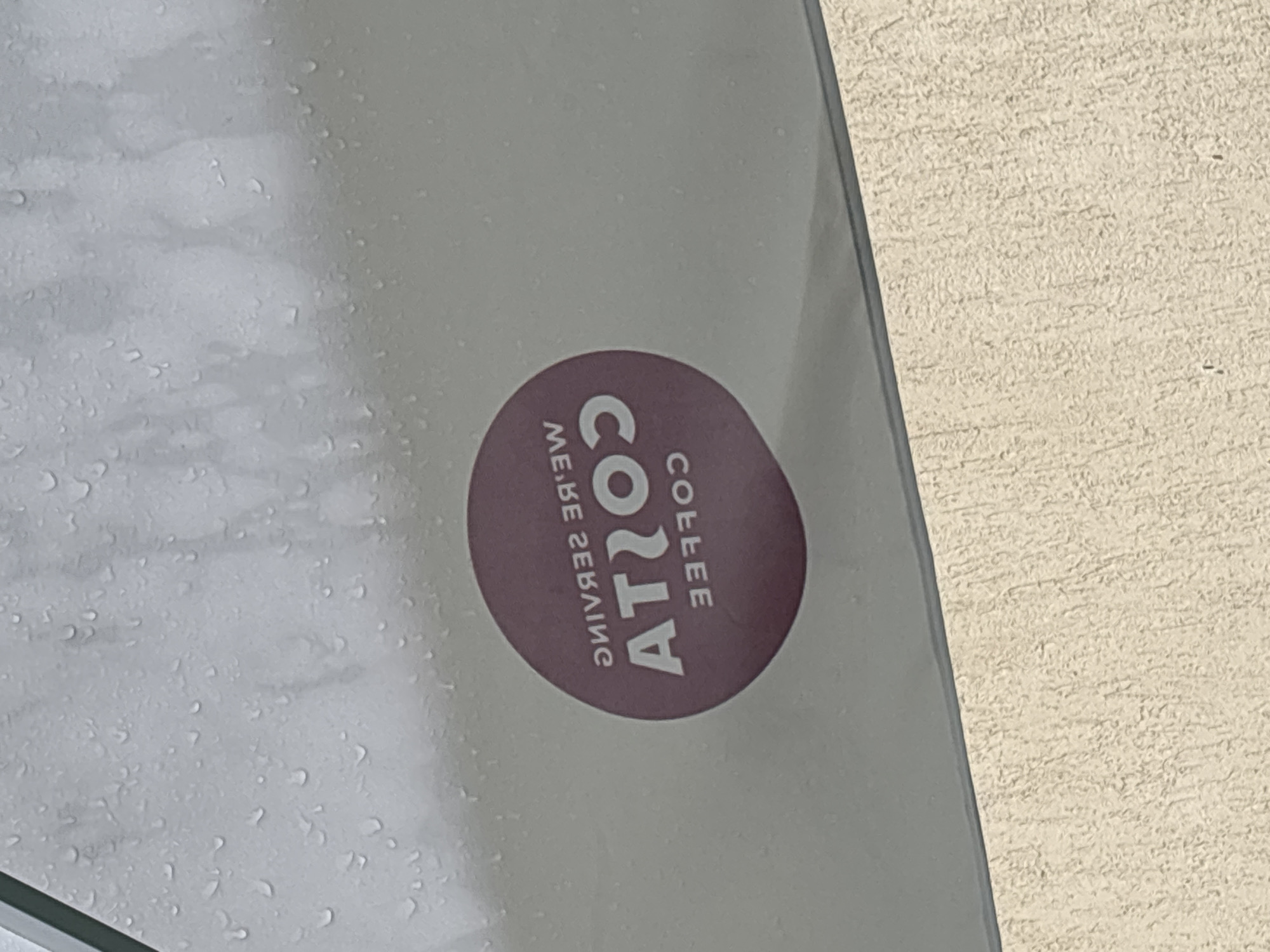 The image captures a close-up, upward-angled view of a wet, light gray surface, possibly an awning or window, adorned with numerous water droplets. A circular, reddish-purple emblem is visible on this surface. The text on the emblem is mirrored; when un-mirrored, it reads "WE'RE SERVING" across the top, "COSTA" in large letters centrally, and "COFFEE" below. The lower section of the image shows a textured, light beige surface, indicative of a wall or ground. The scene is located in Garmen, Bulgaria.