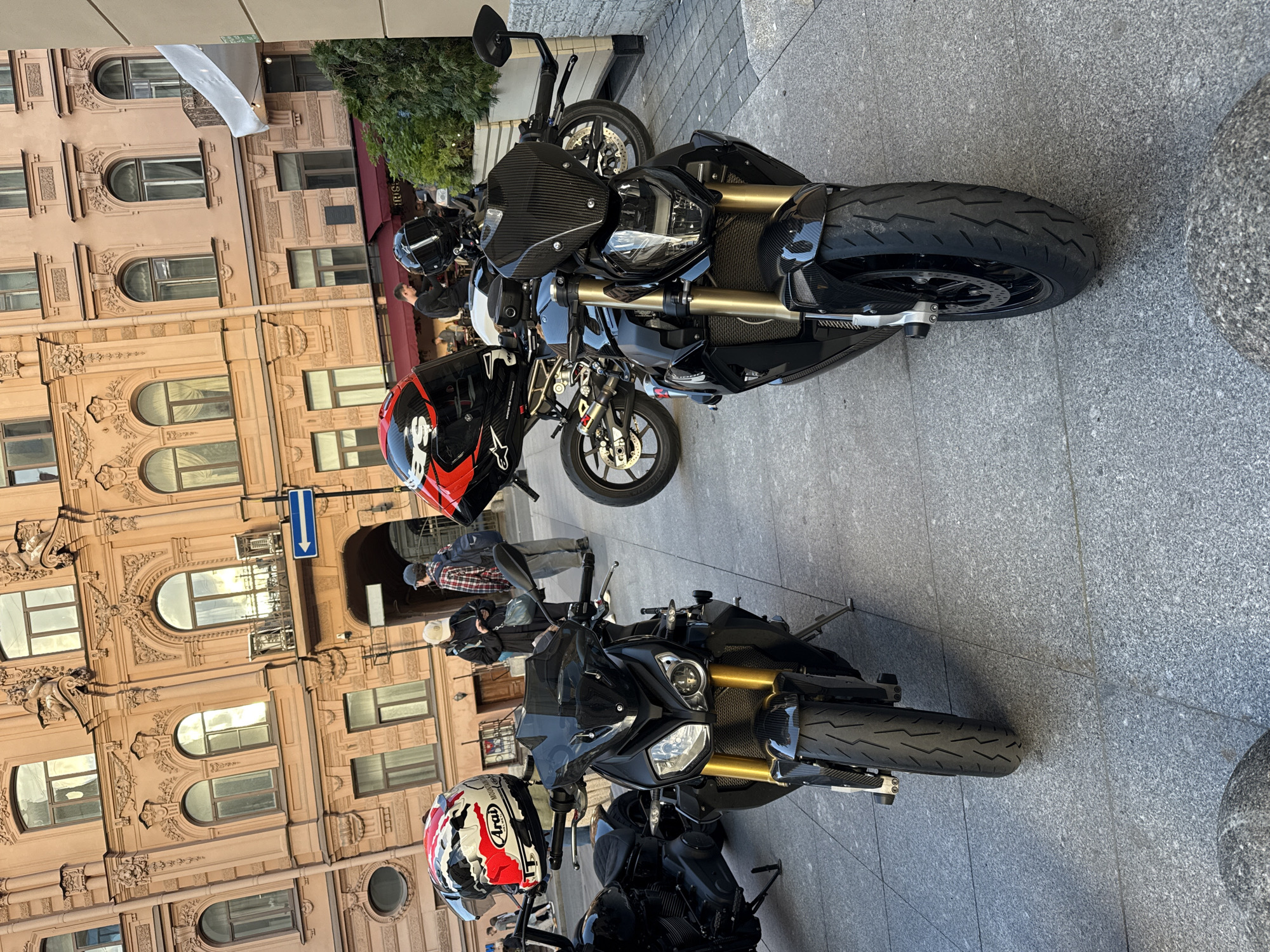 The image shows two black sport motorcycles parked on a sidewalk in front of an ornate building in Saint Petersburg, Russia. The building, likely a historic structure, features elaborate architectural details such as arched windows, balconies, and decorative moldings, painted in a warm, light brown hue. Sunlight reflects off the windows, suggesting a daytime scene.
On the left motorcycle, an Arai helmet with a red, white, and black design is placed on the handlebar. The motorcycle itself has gold-colored front suspension components and a carbon fiber fender. In the background, two individuals are visible, one wearing a light-colored cap and the other a plaid shirt and backpack. A blue directional arrow sign is mounted on the building.
The second motorcycle, positioned to the right and slightly behind the first, is also black with visible carbon fiber accents on its fairing. It has a clear windshield. A black helmet is resting on its handlebars. The ground is paved with gray granite tiles. Green foliage from an outdoor seating area with a red awning is visible to the right of the motorcycles. The overall scene depicts a street in a European city, possibly a popular tourist or commercial area given the architecture and the presence of parked motorcycles.