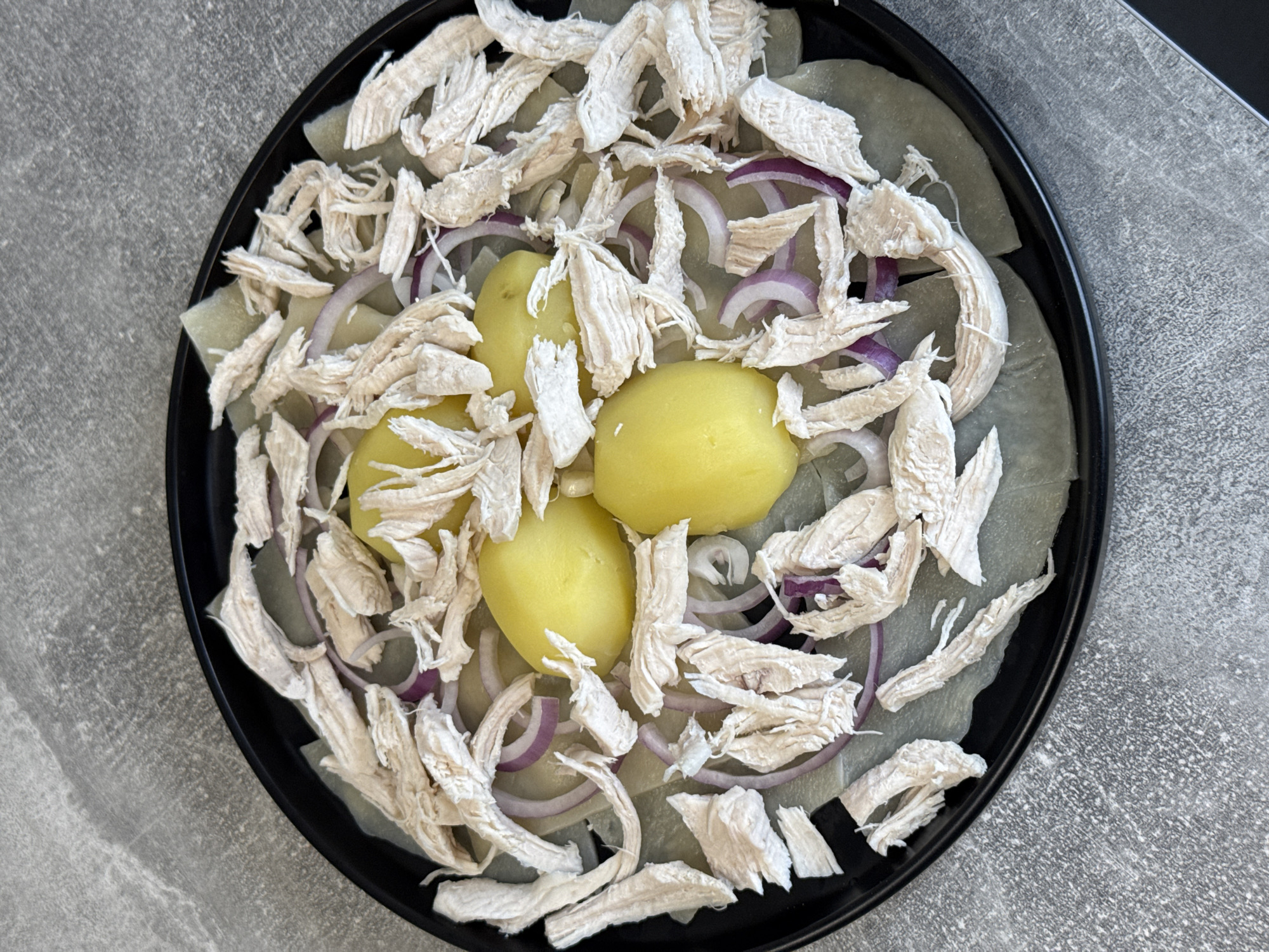 In Lyubertsy, Russia, on a grey, textured surface, a black circular plate holds a dish. The plate contains thinly sliced, pale green vegetables as a base, layered with thin slices of red onion. Four boiled, yellow potatoes are arranged in the center. Shredded, cooked chicken breast is generously piled on top of the potatoes and onions, with some pieces also scattered around the edges of the plate. The lighting is overhead, casting minimal shadows.