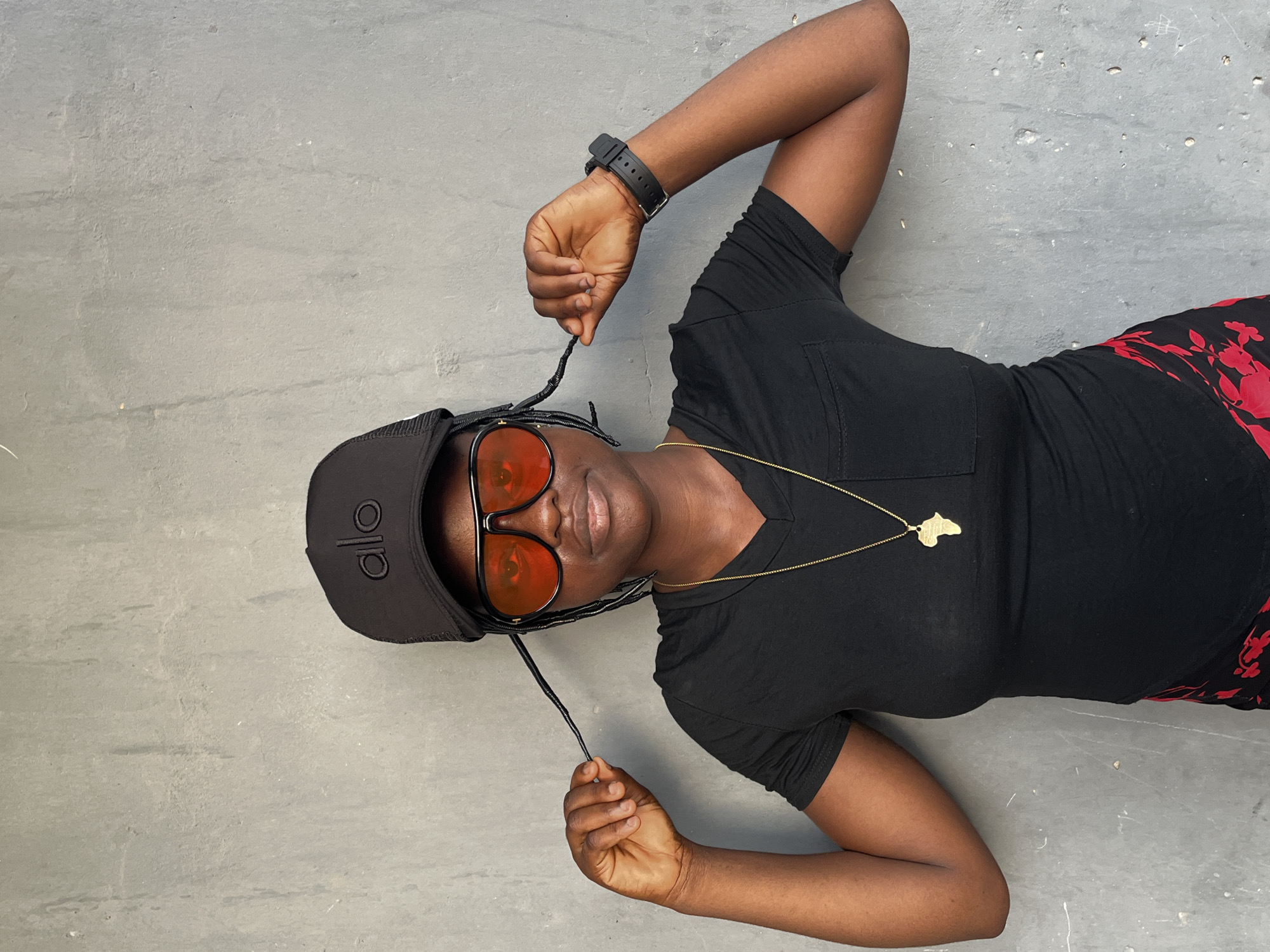 A dark-skinned woman lies on a textured gray surface. She wears a black t-shirt with a v-neck, a black cap with the word "alo" on it, and orange-tinted sunglasses. She also has a gold necklace with an Africa-shaped pendant. She is holding the strings of her cap in both hands. She is also wearing a black watch. Her pants are black with a red floral print. The image does not provide any information about the location in the city of Badagry, Nigeria.