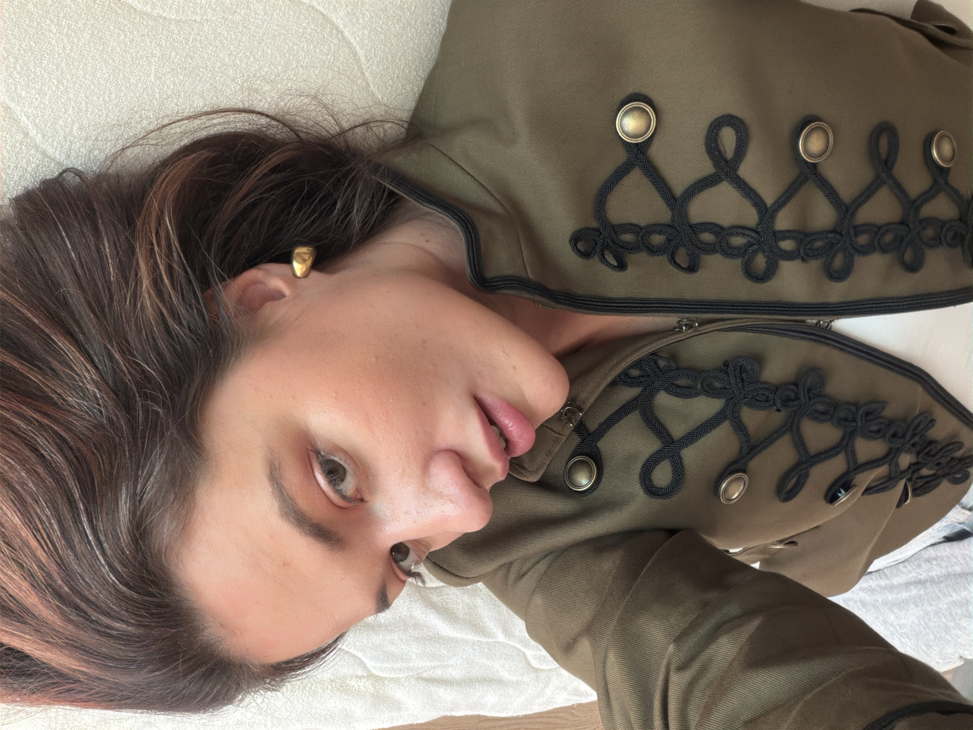 A woman is lying on a white textured surface, possibly a bed, wearing a military-style olive-green jacket adorned with black braided trim and metallic buttons. Her head rests on the surface, and her hair, a mix of brown and highlights, falls around her face. She has a gold stud earring in her ear. The background appears blurred, suggesting a shallow depth of field. The location in Belgrade, Serbia cannot be confirmed from the image alone.