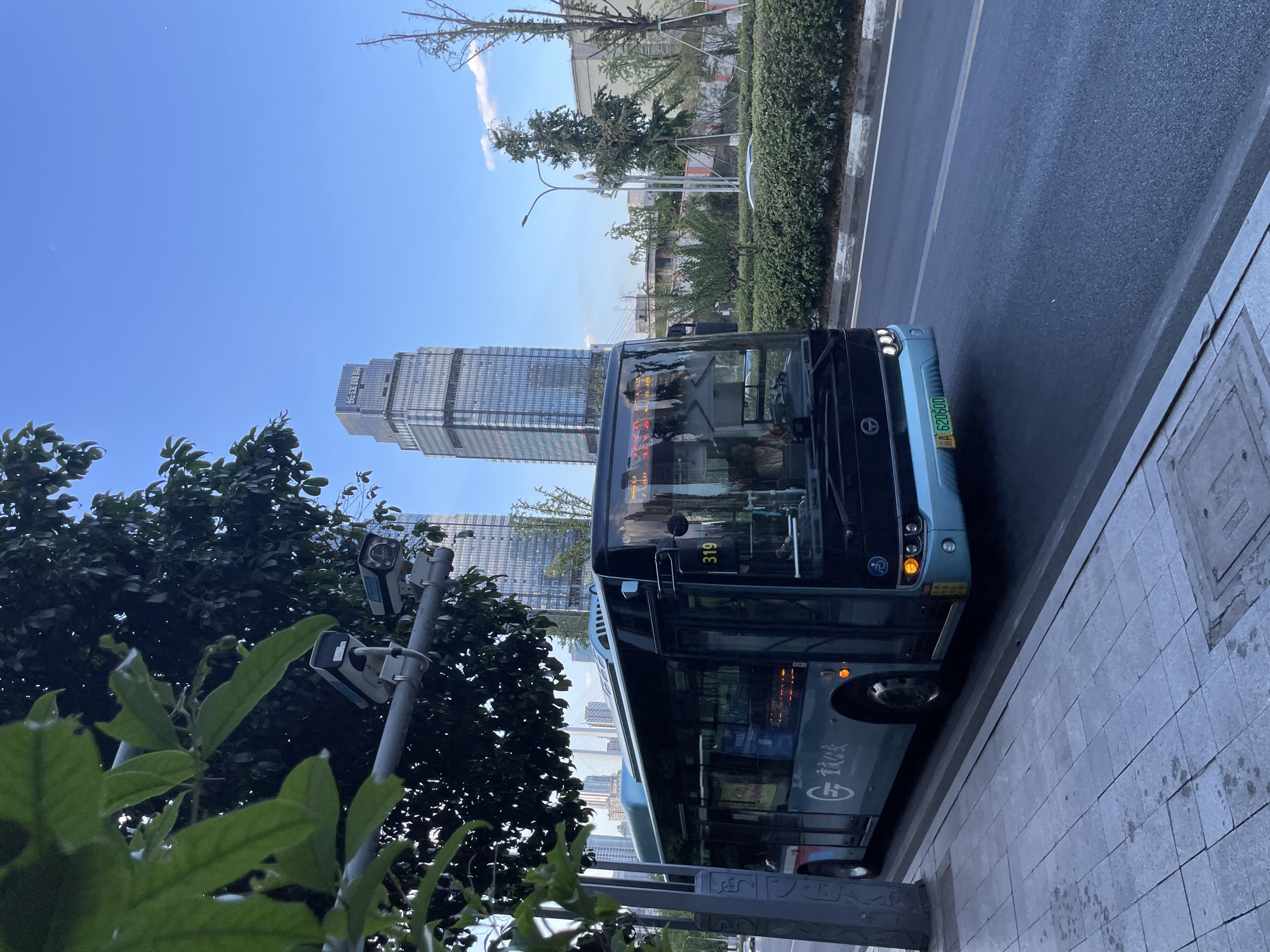 A light blue city bus, displaying the number "319" and the license plate "A 62060D", is parked alongside a paved sidewalk. The bus has black-tinted windows and visible interior lights. Two surveillance cameras are mounted on a pole above the sidewalk, partially obscured by green foliage. In the background, two modern skyscrapers with glass facades pierce a clear blue sky. A bridge can be seen in the distance between the buildings, and mature trees and manicured hedges line the street. The scene appears to be a well-maintained urban environment, likely in the Nan'an District of China.