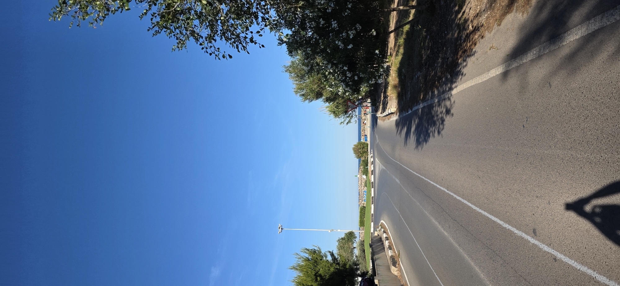 The image depicts a road in Canet d'en Berenguer, Spain. The road curves slightly, with a white line marking its edge. Vegetation, including trees and bushes, lines one side of the road. In the distance, there is a view of the beach and the sea. The sky is clear and blue. The scene appears to be captured during the daytime, with shadows cast by trees along the road. People are faintly visible in the distance, near the beach. A lamppost stands adjacent to the road. The overall impression is a sunny coastal road scene.