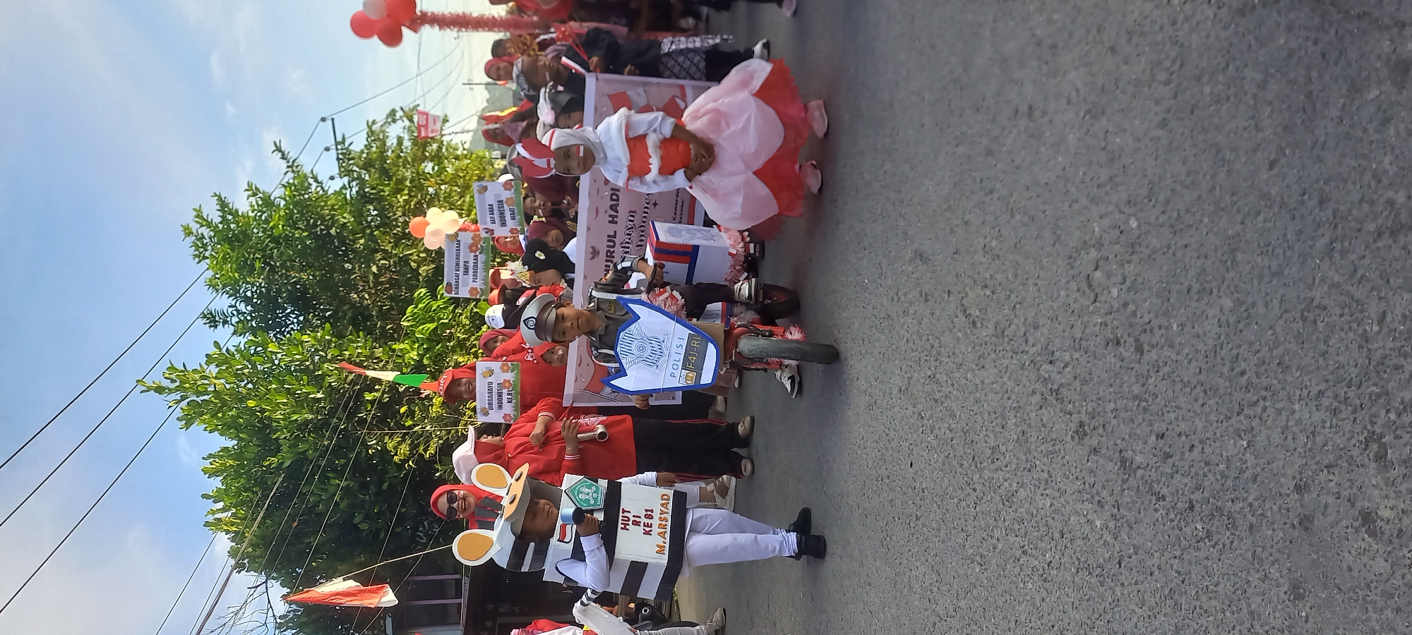 This is a medium shot, outdoor, eye-level, vertical photograph taken on a paved street in Samarinda, Indonesia. The scene depicts a parade or procession, likely celebrating Indonesian Independence Day given the presence of red and white flags and banners.
In the foreground, two children are the primary subjects. The child on the left is dressed in a black and white striped costume with large, round ears, resembling a character like a bee or a prisoner. The child is holding a microphone and has a sign on their costume that reads "HUT RI KE 81 M. ARSYAD". The child on the right is dressed as a police officer, wearing a uniform and a cap, and is seated on a small, red toy motorcycle. This child is holding a shield with the word "POLISI" and a license plate "F4J-RI" on it. Behind these two children, other children and adults are visible, many wearing red and white attire. Several banners are held up by adults, some displaying text in Indonesian. The background shows trees, power lines, and a clear blue sky with some clouds. The overall atmosphere appears festive and celebratory.