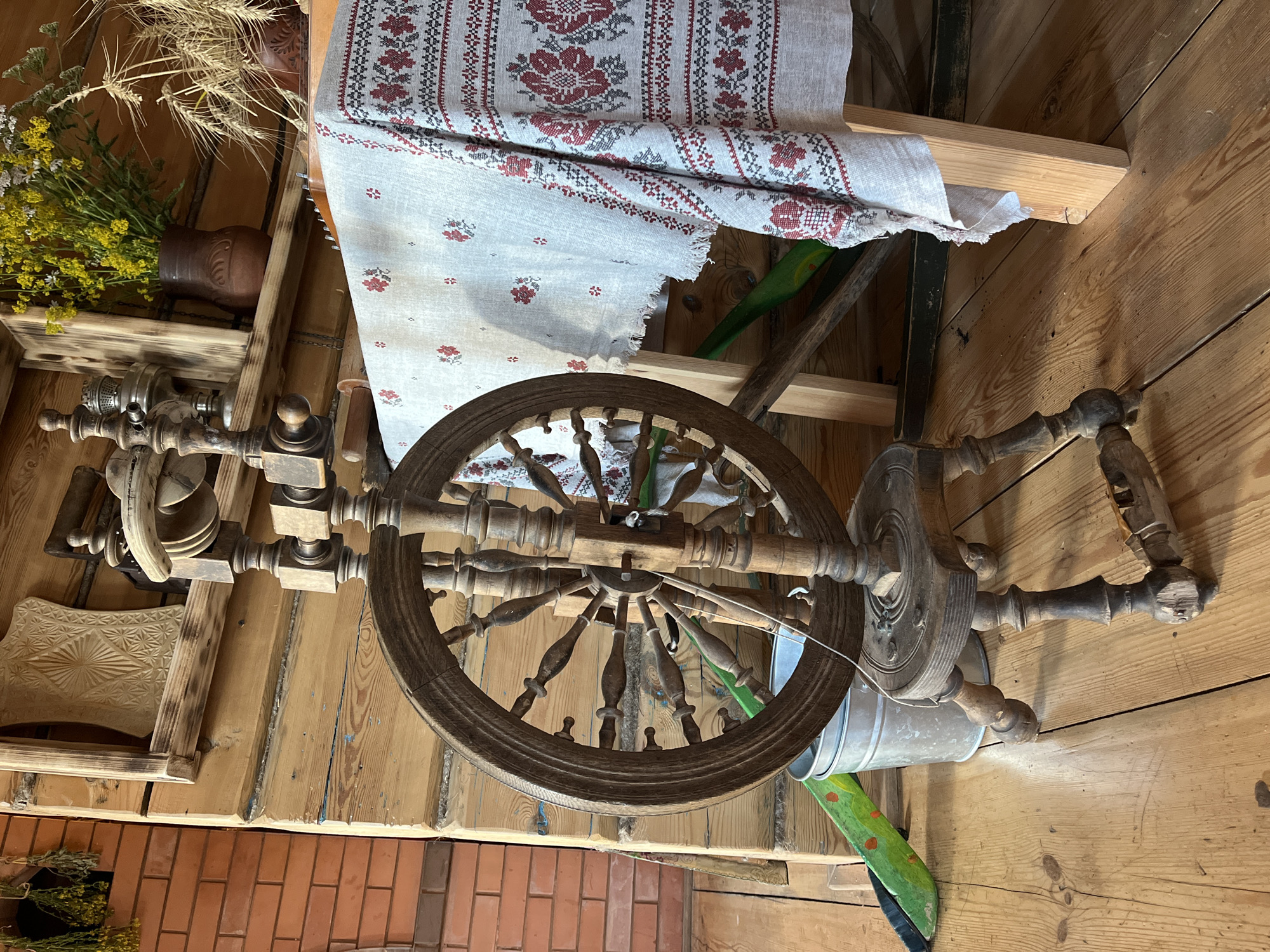 A close-up, vertically oriented medium shot captures a spinning wheel and a piece of embroidered fabric in a rustic, indoor setting. The spinning wheel, made of aged, dark wood, is the central object, with its large wheel and intricate details visible. To the upper right of the spinning wheel, a light-colored fabric with red and grey embroidered floral patterns is draped over a wooden structure, suggesting a workshop or display area.
In the background, wooden plank walls and shelves are visible, adding to the rustic ambiance. On a shelf to the upper left, a clay jug sits next to a bouquet of dried wheat and yellow wildflowers, hinting at a rural or traditional setting. A patterned, light-colored object with intricate carved details is placed on a wooden shelf below the spinning wheel. A galvanized metal bucket is also visible near the base of the spinning wheel.
The overall impression is one of traditional craftsmanship and a connection to rural life in Russia, possibly in Vybolovo given the location context. The lighting appears to be natural, coming from an unseen source, and the overall mood is one of quiet history and handmade artistry. There is no visible text in the image.