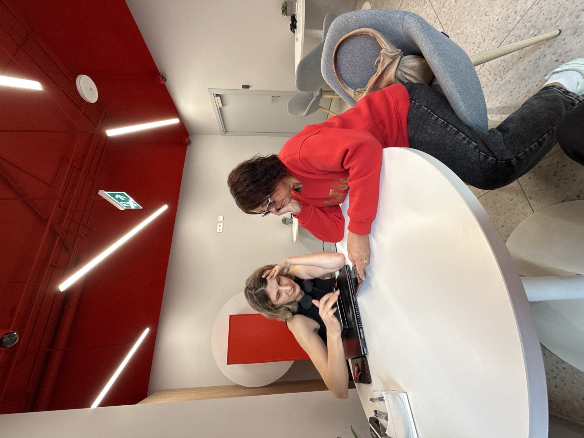 In Zheleznogorsk, Russia, two women are seated at a round white table. The woman on the left, with blonde hair, wears a black sleeveless top and has headphones around her neck. She is looking down at a laptop and a phone on the table, with her left hand resting on her forehead. The woman on the right, with dark hair, wears a red sweatshirt and dark jeans. She is wearing glasses and looking at a phone. A light brown purse is draped over the back of her gray chair. The environment features a red ceiling with modern lighting fixtures and an exit sign. The walls are light gray.