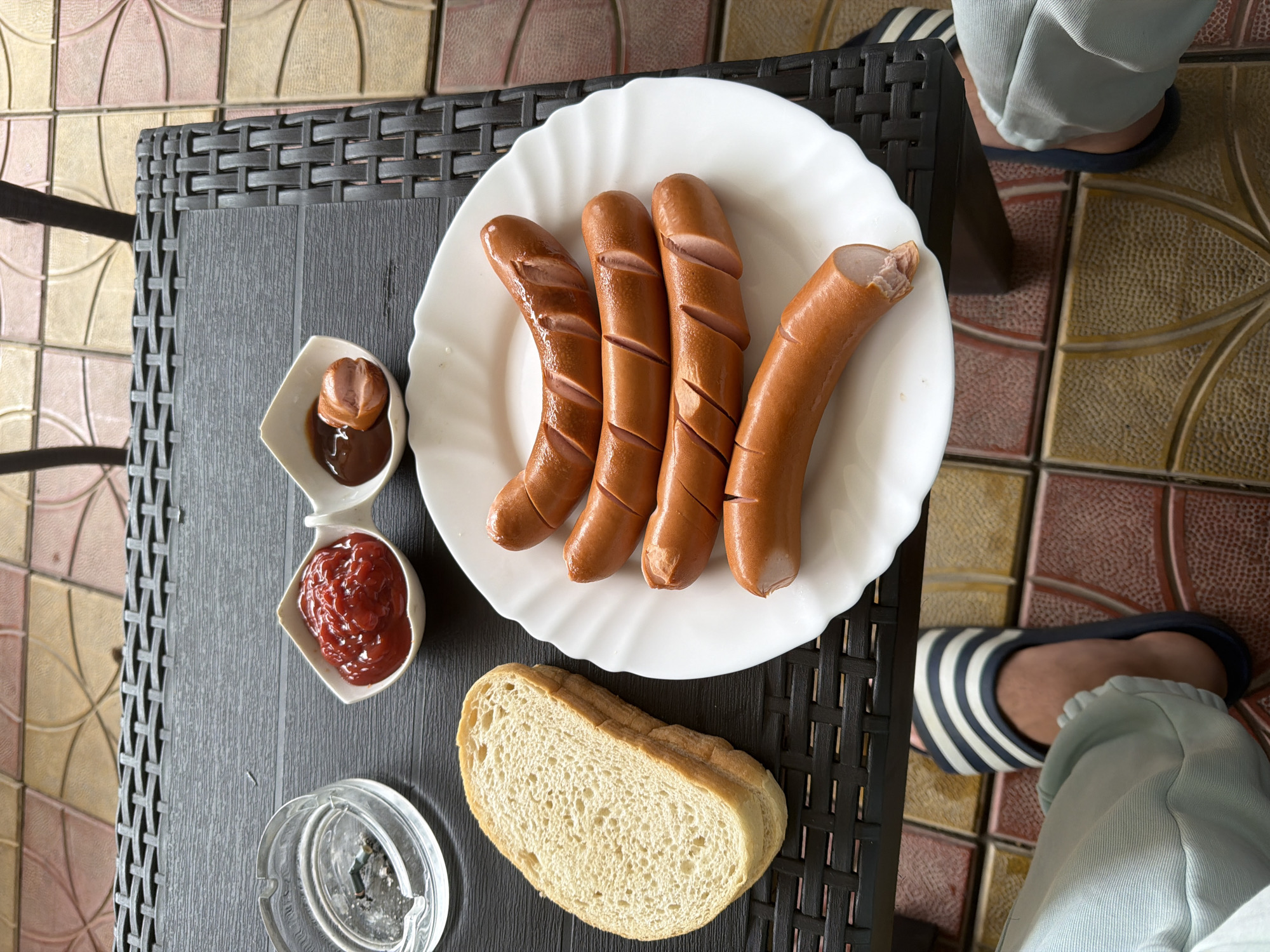 An overhead shot captures a simple meal laid out on a dark grey, woven-patterned table, likely on an outdoor patio or balcony in Dragor, Bulgaria. The main focus is a white scalloped plate holding five cooked hot dogs or sausages. Four of the sausages exhibit diagonal cuts along their length, while one on the far right has a bite taken from its end, revealing the light pink interior.
Adjacent to the plate are two small white, heart-shaped dishes containing condiments. One dish holds a dollop of red ketchup, and the other contains a darker, brown sauce (possibly BBQ sauce or mustard) with a small, cut piece of sausage submerged within it. A thick slice of white bread rests on the table next to the condiments.
In the bottom left corner of the frame, a clear glass ashtray with some ash is visible. The lower right portion of the image shows the legs and feet of a person, dressed in light blue casual pants and wearing black-and-white striped flip-flops or sandals. The floor beneath the table is adorned with decorative, multi-toned tiles featuring geometric patterns in shades of brown, beige, and pink. The lighting is bright and even, suggesting daytime. No visible text is present in the image.