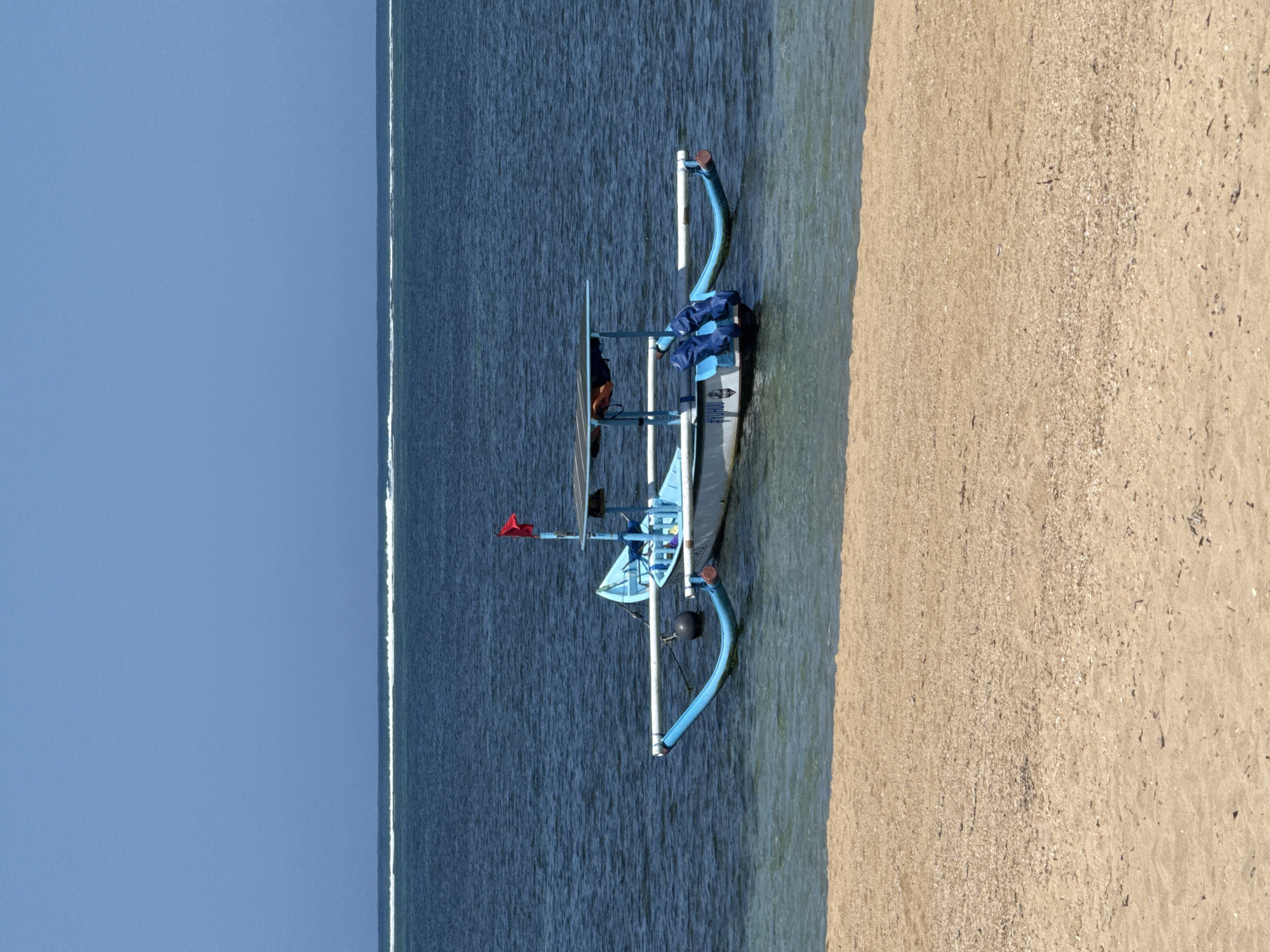 A traditional Balinese outrigger boat, painted blue, rests on the shallow water near the sandy shore of Sanur Beach in Denpasar, Indonesia. The boat features a canopy providing shade and is equipped with two outriggers extending to the sides for stability. A small red flag is attached to a pole at the stern. The clear blue sky forms the backdrop, with a faint white line of distant waves visible on the horizon. The foreground is dominated by the tan sand of the beach, sparsely scattered with small shells and debris. The water is a deep blue, transitioning to a lighter, clearer hue near the shore. No people are visible in the image.