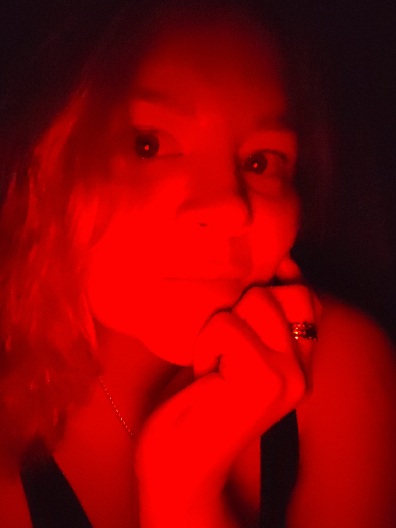 A close-up, dimly lit portrait of a woman with her hand resting on her chin. The entire image is cast in a strong red light, making it difficult to discern details. She is wearing what appears to be a ring on one of her fingers and a chain around her neck. No evidence suggests that this image was taken in Izhevsk, Russia.