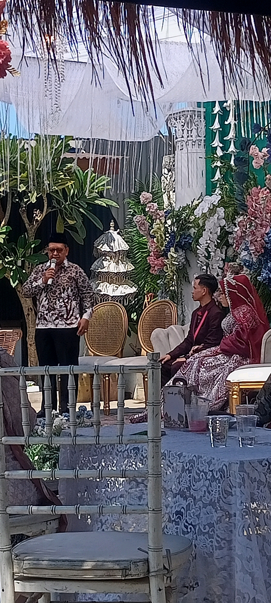 A man wearing a patterned shirt and black pants stands at a podium, holding a microphone and speaking. He is on a raised platform decorated with hanging fabric and foliage, with a tiered decorative item to his right. Seated on the platform to the right of the speaker are a man and a woman. The man is wearing a maroon jacket and the woman is wearing a red wedding dress and veil. They are seated on chairs with woven backs. A table covered with a white lace cloth is in front of the seated couple. The scene appears to be an outdoor wedding ceremony in Desa Ponokawan, Indonesia.