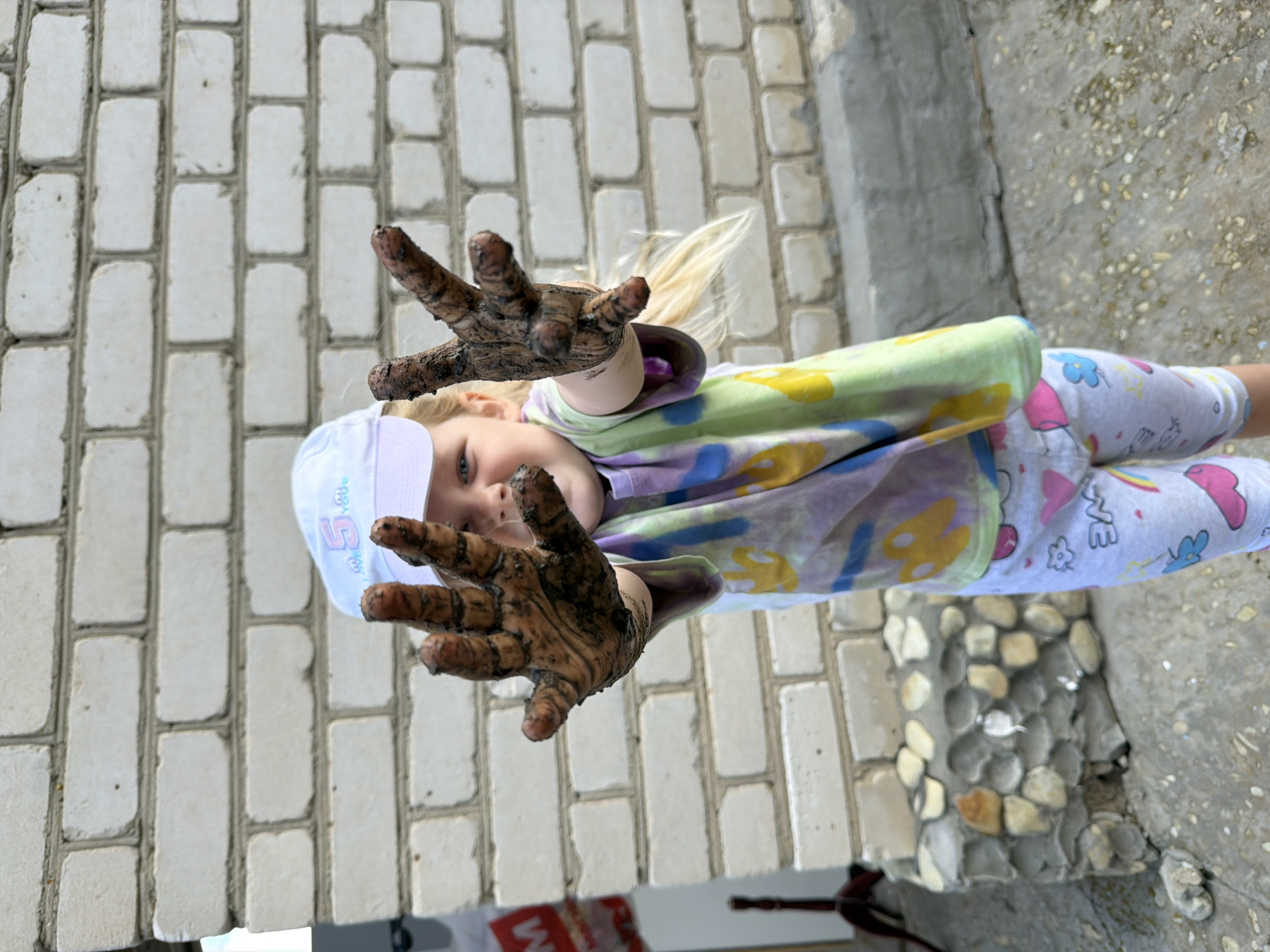 The image captures a young girl with blonde hair, wearing a white cap and a tie-dye t-shirt with a colorful design, standing in front of a white brick wall. Her hands are covered in dirt, and she is holding them up, displaying the mud. She is wearing patterned leggings. In the background, a portion of a concrete structure with embedded stones is visible. The scene appears to be outdoors, possibly in a yard or a public space in the city of Kamyshin, Russia, given the context of the prompt. There is a red and white sign on the left side of the image.