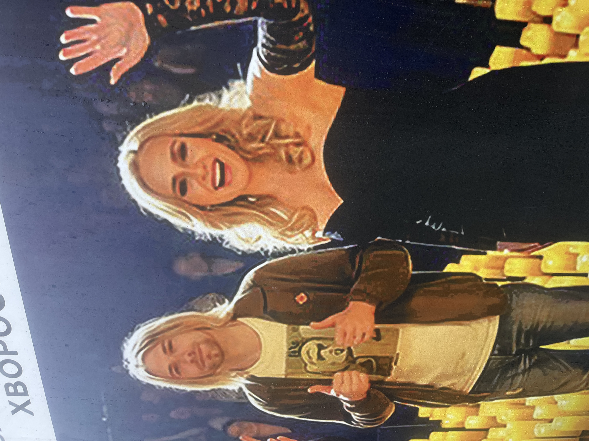 This image appears to be a close-up of a digital screen or a print, displaying a photograph or artwork featuring two celebrities. At the top is a woman with blonde curly hair, wearing a black dress with lace sleeves. She is smiling with her mouth open and her right hand is raised. Below her is a man with long blonde hair and a brown jacket over a white t-shirt with a graphic on it. He is making a thumbs-up gesture with both hands. Both people are superimposed against a background of what appears to be small gold bars. The image is tilted and partially obscured by what appears to be white lettering on the left edge, reading "XBOPO" vertically. No specific location within Krasnoyarsk, Russia, is identifiable from this image.
