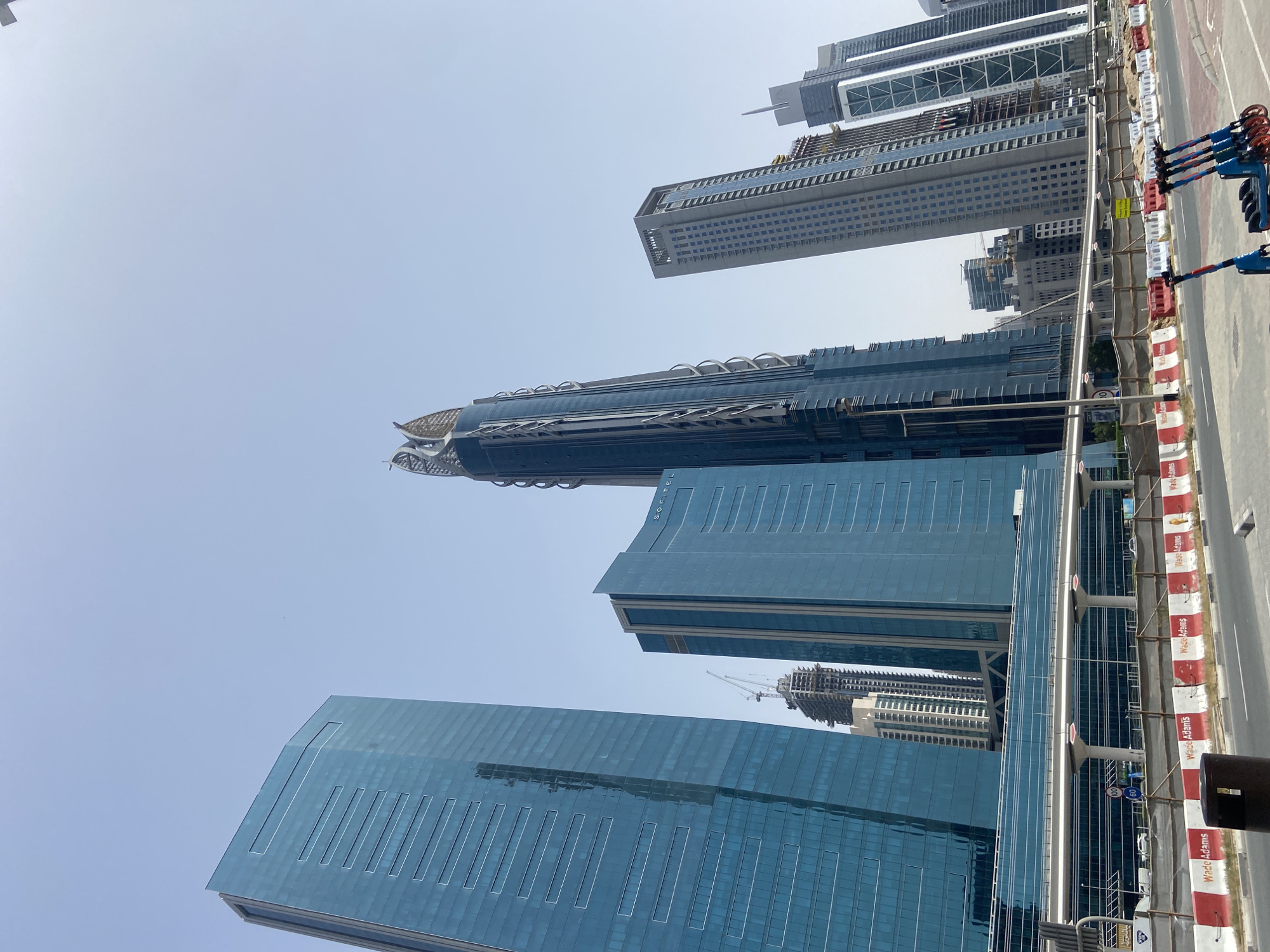 The image captures a view of Dubai's skyline in the United Arab Emirates. Several modern skyscrapers dominate the frame, showcasing a blend of architectural styles. The buildings are clad in reflective glass and metal, reflecting the sky. A construction crane is visible among some of the buildings, indicating ongoing development in the area. Adjacent to the buildings is a street with barriers and signage, which seems to be part of a construction or roadwork setup. The sky is a clear blue, suggesting a sunny day. The angle is a low-angle shot, giving a sense of the buildings' height.