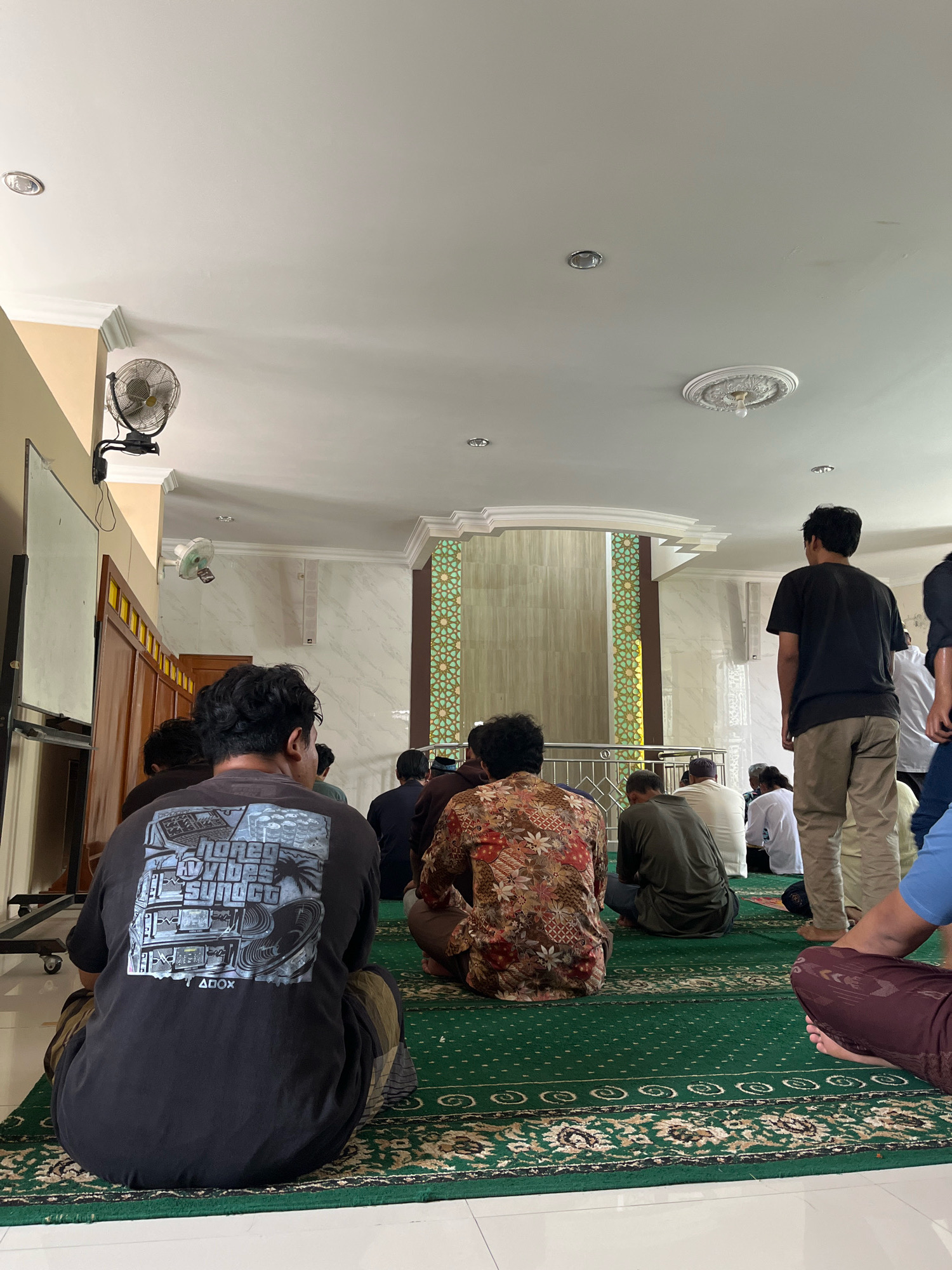 The image depicts the interior of a mosque in Gentan, Indonesia, during a prayer service. Several men are seated on a green carpeted floor, facing towards a decorative mihrab. In the foreground, a man wearing a black t-shirt with a graphic print is seated, facing away from the camera. To his right, other men are also seated or standing, some in prayer attire. The mosque has white walls and a high ceiling with decorative molding and lighting fixtures. A large fan is mounted on the wall on the left side of the image. The overall environment suggests a peaceful and communal gathering for religious observance.