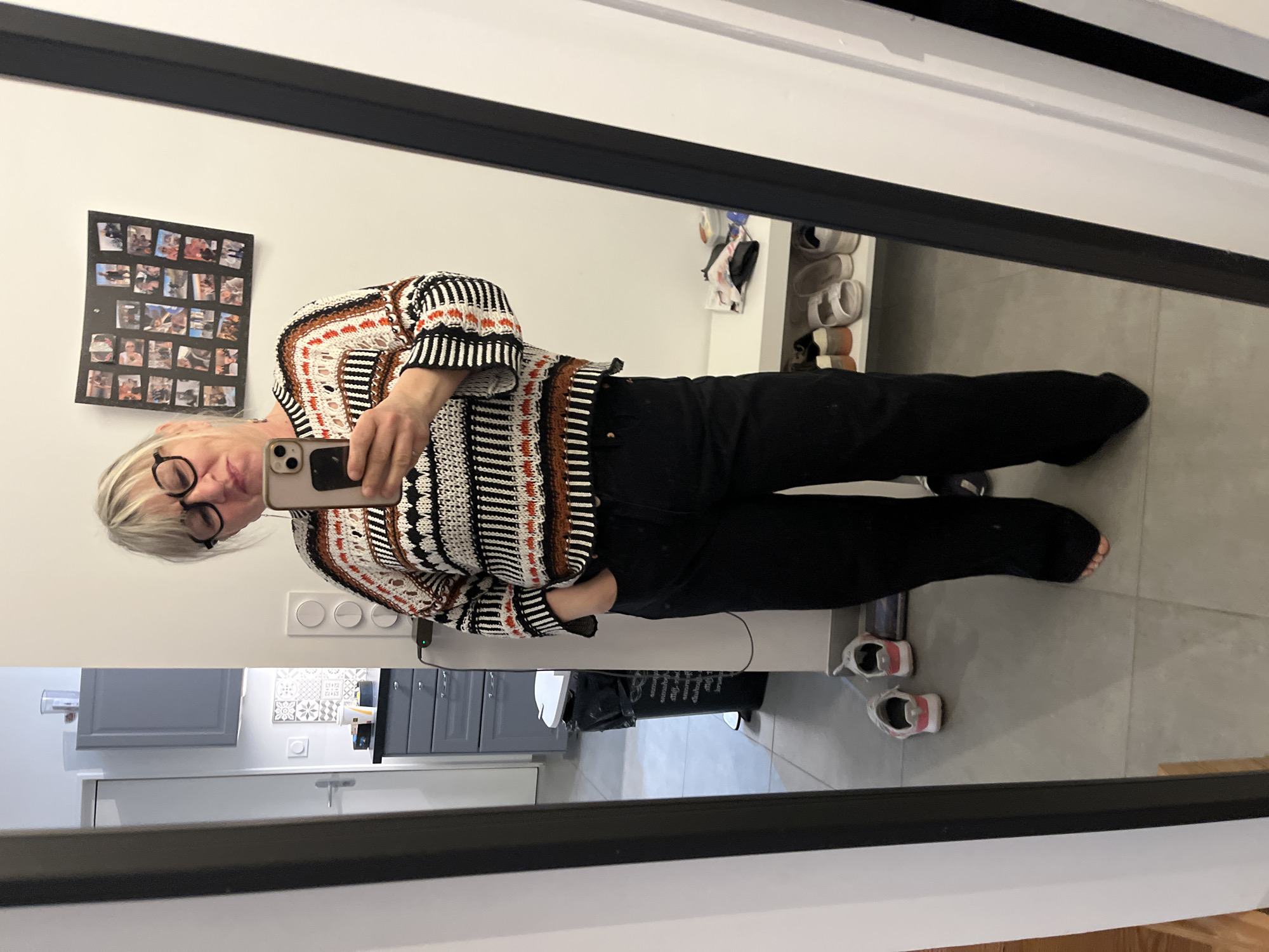 A woman is reflected in a mirror, presumably taking a selfie with a smartphone. She is wearing glasses, a striped sweater with orange accents, and black pants. Behind her, a wall displays a photo collage. In the background, there is a shoe rack filled with shoes, a gray kitchen area with cabinets, a white countertop, and a trash bag. The setting appears to be an indoor residential space. There is no indication of the location of Chambœuf, France.