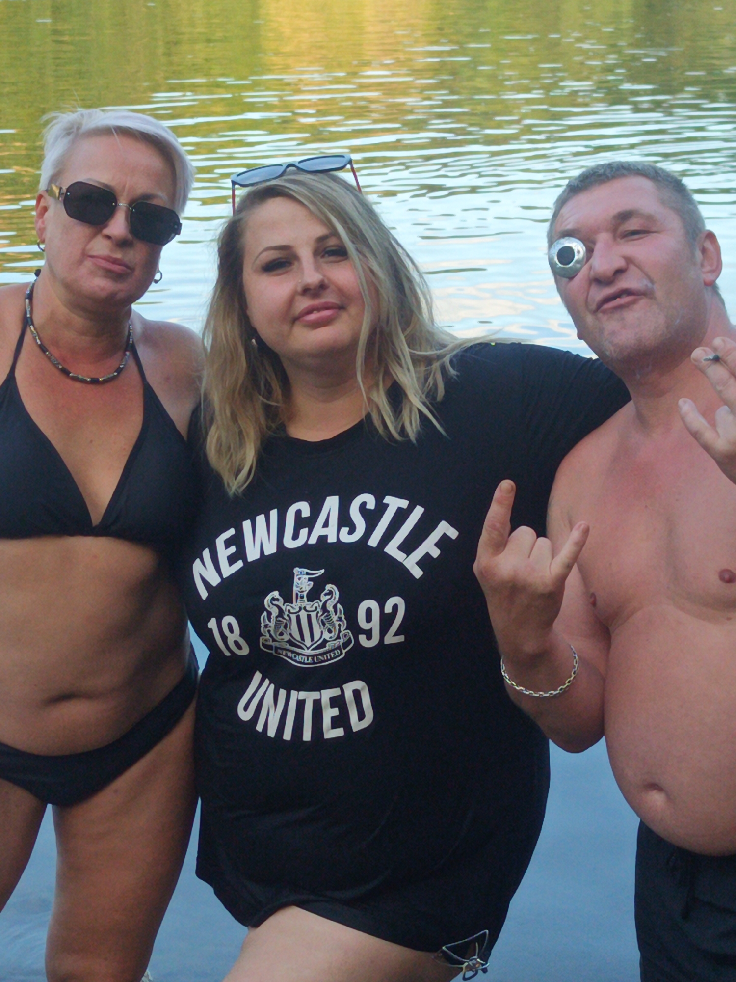 Three individuals are posing for a photo in shallow water with a body of water in the background. On the left is a woman with short blonde hair, wearing sunglasses and a black bikini top. In the center is a woman with blonde hair, wearing a black t-shirt with "Newcastle United 1892" printed on it. On the right is a man with short grey hair, wearing no shirt, with a silver object over his right eye. He is making a "rock on" hand gesture. The man on the right is also smoking a cigarette. The water is calm and appears to be a lake or river. The background shows a blurred green and yellow reflection, suggesting foliage or a distant shore. There is no indication of the specific location within Uzhhorod, Ukraine.