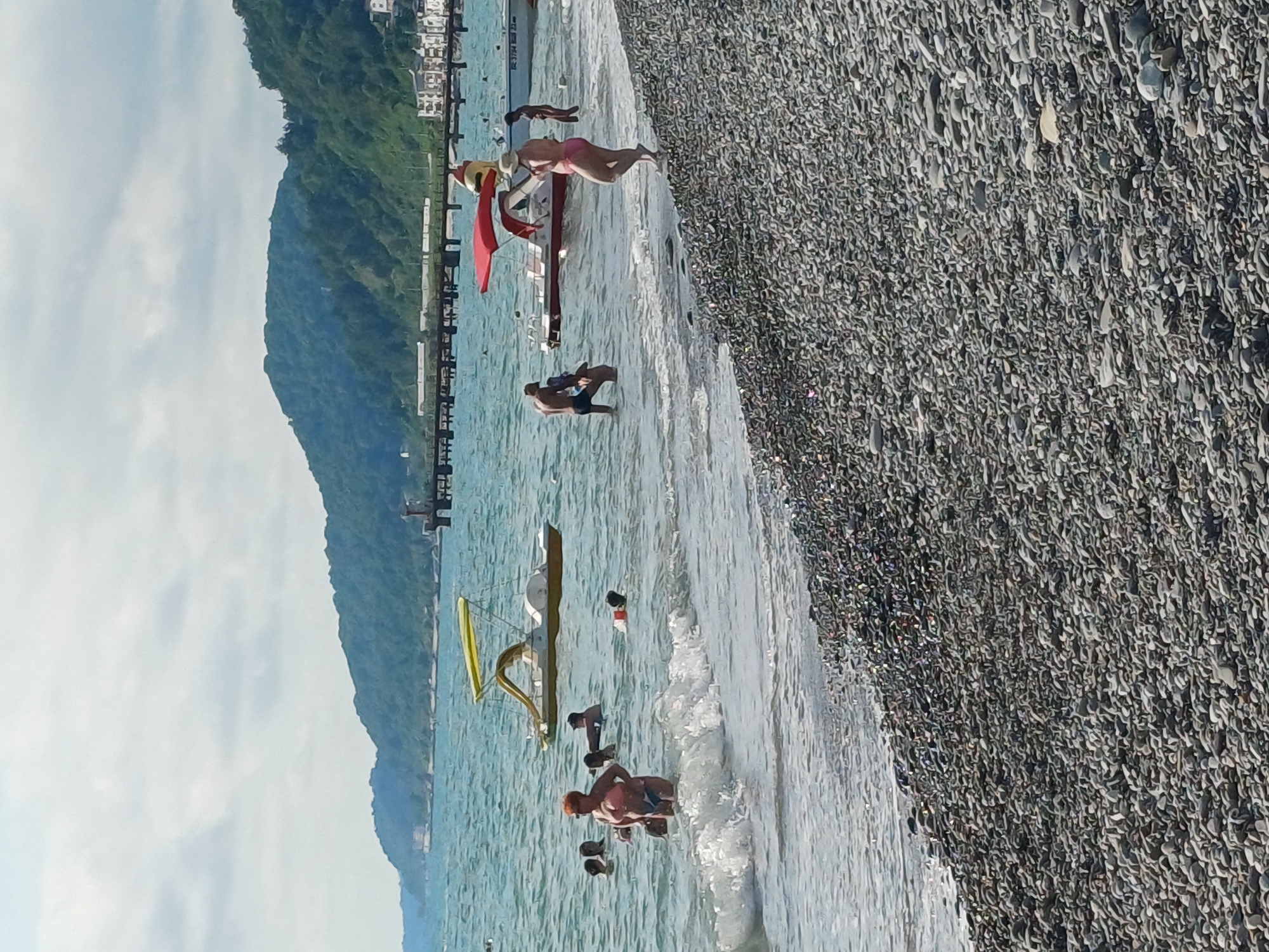 The image depicts a sunny day at a pebble beach in Sochi, Russia. Several people are engaged in recreational activities in the shallow, turquoise waters of the sea. A woman in a pink bikini is seen on a structure in the water, possibly a part of a water slide or play area. Another individual is partially submerged in the water nearby. Further out, two men are interacting in the sea, with one appearing to hold onto the other. Closer to the shore, a group of people are in the water, with one individual in the foreground wearing an orange cap, holding what appears to be a child. The beach itself is covered in a dense layer of grey pebbles. In the background, a lush, green mountain slopes down towards the sea, with buildings visible at its base. A pier or walkway extends into the water. The water is calm with small waves lapping at the shore. The sky is partly cloudy. The overall atmosphere suggests a pleasant summer day at the beach, with people enjoying the water and the scenery. There is no visible text in the image.