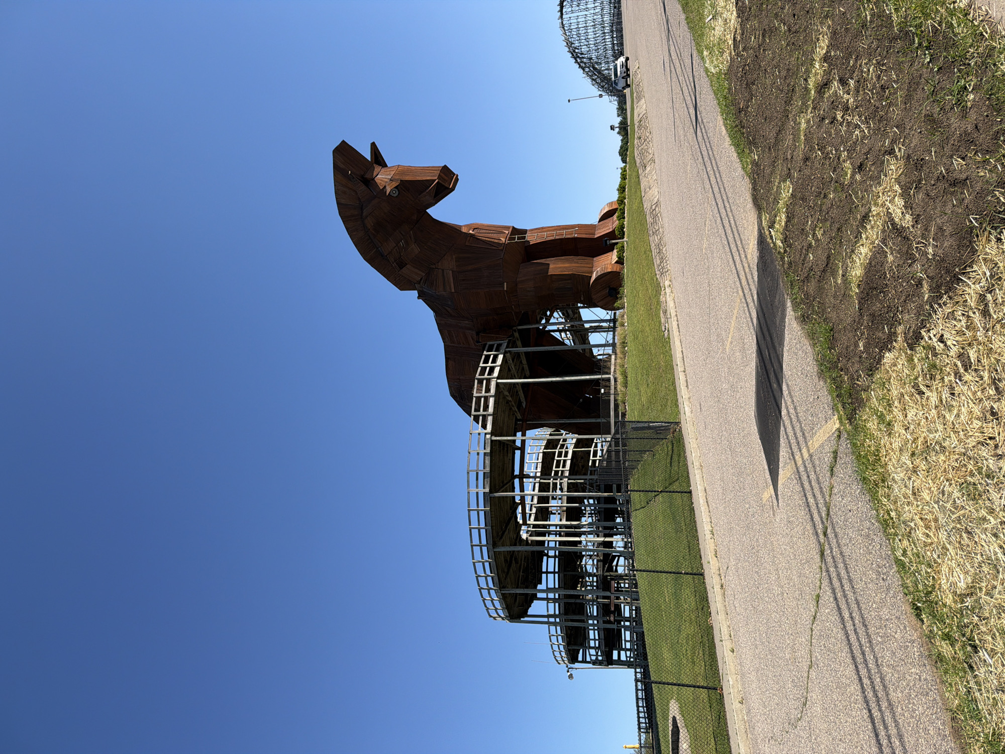 The image shows a large, wooden Trojan horse sculpture located in Lake Delton, Wisconsin. The horse is brown and appears to be constructed from planks of wood. It stands on a metal scaffolding structure, and a black fence surrounds the base. A paved pathway runs alongside the sculpture, with patches of grass and bare earth on the other side. The sky is clear and blue. In the background, a large, silver, dome-shaped structure is visible.