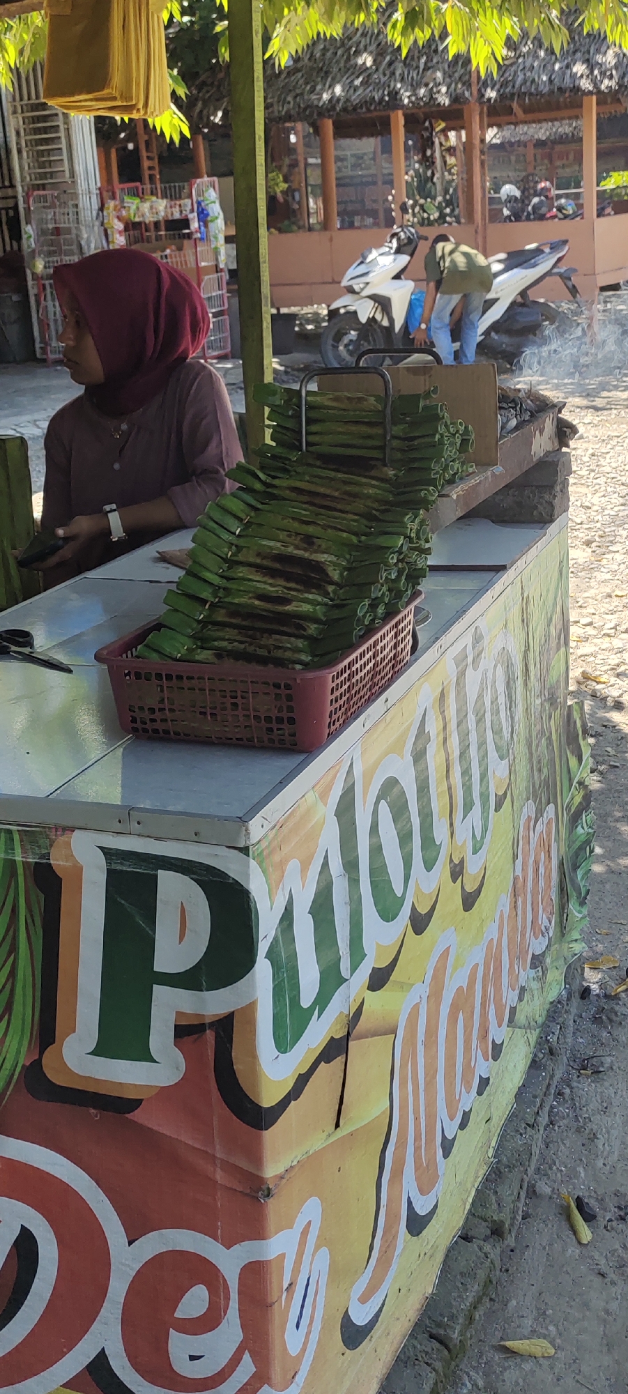 The scene depicts an outdoor food stall, likely in Lhokdagang, Indonesia. A woman wearing a maroon hijab sits behind the stall, seemingly operating it. A basket on the counter contains a stack of food items wrapped in green leaves and slightly charred. A grill sits behind the basket, indicating the food is grilled. The stall has lettering on the side that partially spells out "Pulo". In the background, a man stands near a white motorbike, appearing to be working on it. Smoke rises near the motorbike. The location appears to be near a building with a thatched roof. A cart is visible in the background filled with products.