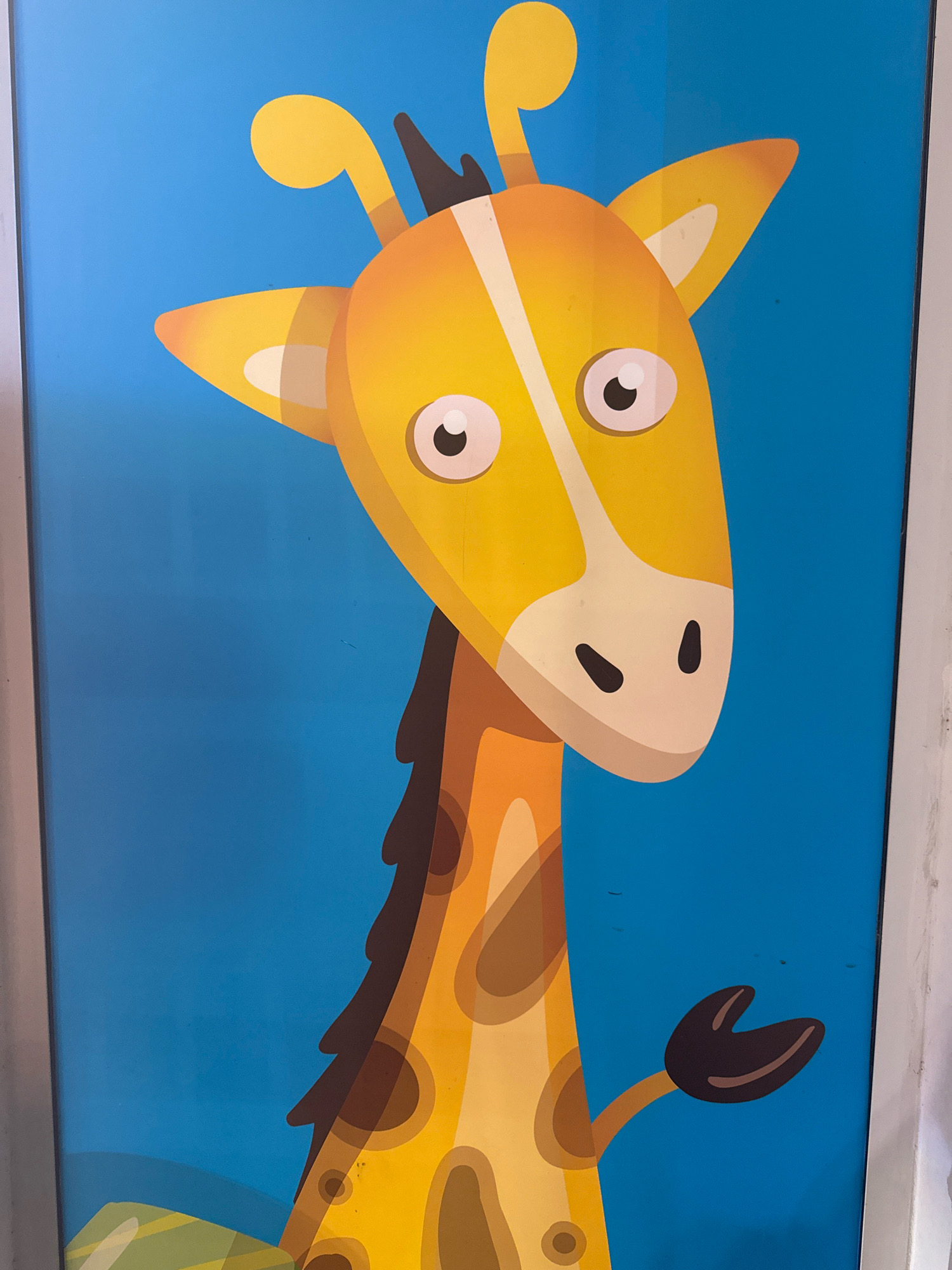 A cartoon giraffe is depicted against a bright blue background. The giraffe's head is large and yellow, with prominent white and black eyes. It has a white snout with two black nostrils and brown spots on its neck. A single dark brown hand-like shape is visible on the right side of the image, extending from the giraffe's body. The giraffe's body is orange with brown spots. The bottom left of the image shows a hint of green and yellow, suggesting grass or foliage. This artwork is located on a wall in Agios Athanasios Municipality, Cyprus.
