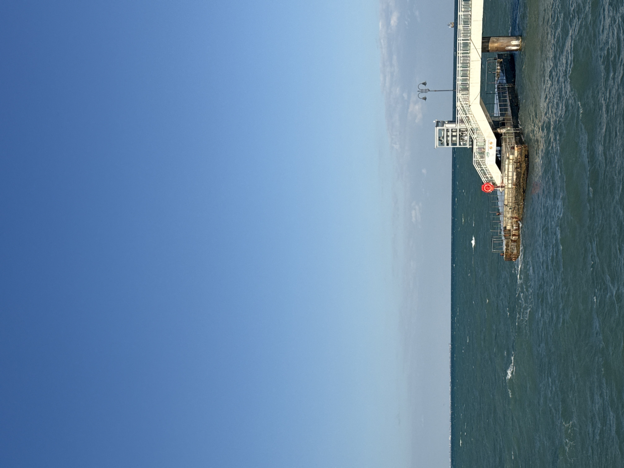 The image depicts a modern pier jutting out into the sea under a clear blue sky. The pier features a white, elevated walkway with railings, accessible by stairs. At the end of the walkway, there is a small white building, possibly a control room or information kiosk, with a vintage-style lamppost nearby. The sea is a deep teal color, with moderate waves indicating a breezy condition. A red circular object, possibly a lifebuoy, is attached to the railing on the left side of the pier. The structure appears to be part of the Burgas Sea Station complex in Burgas, Bulgaria, a popular destination for both locals and tourists, offering recreational and functional facilities by the coast. The scene is serene and orderly, capturing a typical day at the seaside.